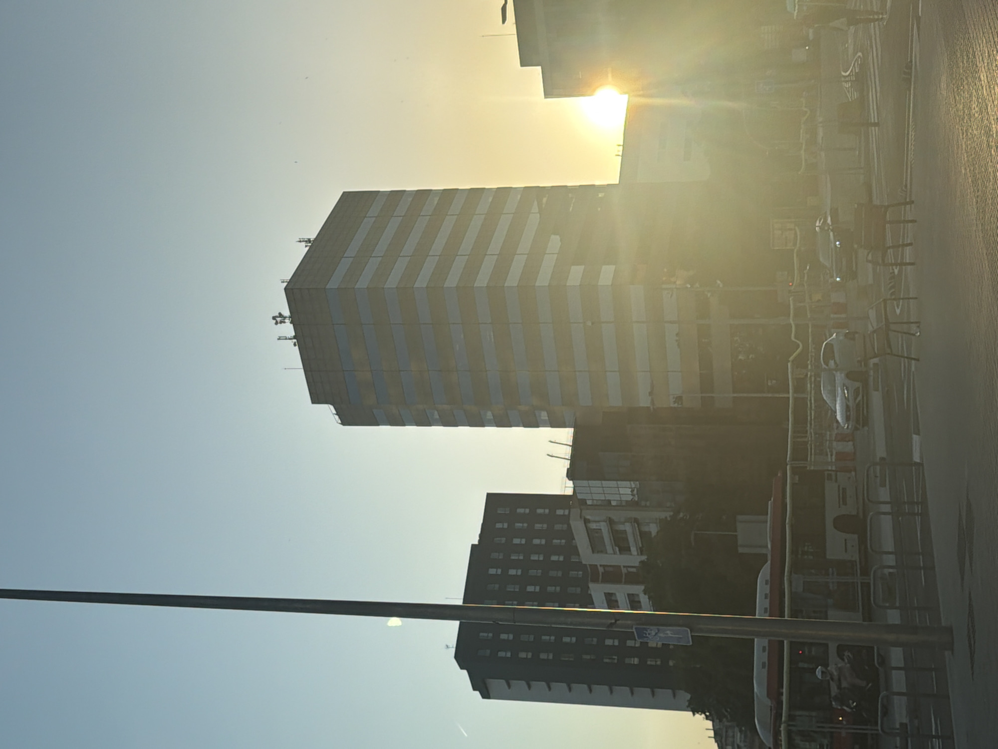 This image depicts a street scene in Barcelona, Spain, at sunset. The golden light of the setting sun creates a strong lens flare in the upper right portion of the frame, illuminating the sky and casting a warm glow over the urban landscape.
Dominating the scene are several tall buildings. The most prominent is a modern skyscraper with a facade of alternating light and dark vertical stripes, likely windows and building materials. To its right, and further in the background, are other buildings, some appearing to be residential blocks with numerous windows. In the foreground, a dark, multi-story building with many visible windows is partially obscured by a metal pole, possibly a street light or traffic signal.
The street itself is visible on the right side of the image, with cars and what appears to be a bus. The presence of bicycles parked in racks suggests a vibrant urban environment. The activity appears to be a normal evening in a city, with no specific event or dramatic action taking place.
Notable details include the time of day, indicated by the sunset and the quality of light. The weather appears clear, with a bright sky. The presence of distinct architectural styles and the street-level details, like the parked bicycles and traffic, are characteristic of a European city. There is a blue and white traffic sign on the metal pole in the foreground. The overall mood is one of a serene urban evening bathed in warm, fading sunlight.