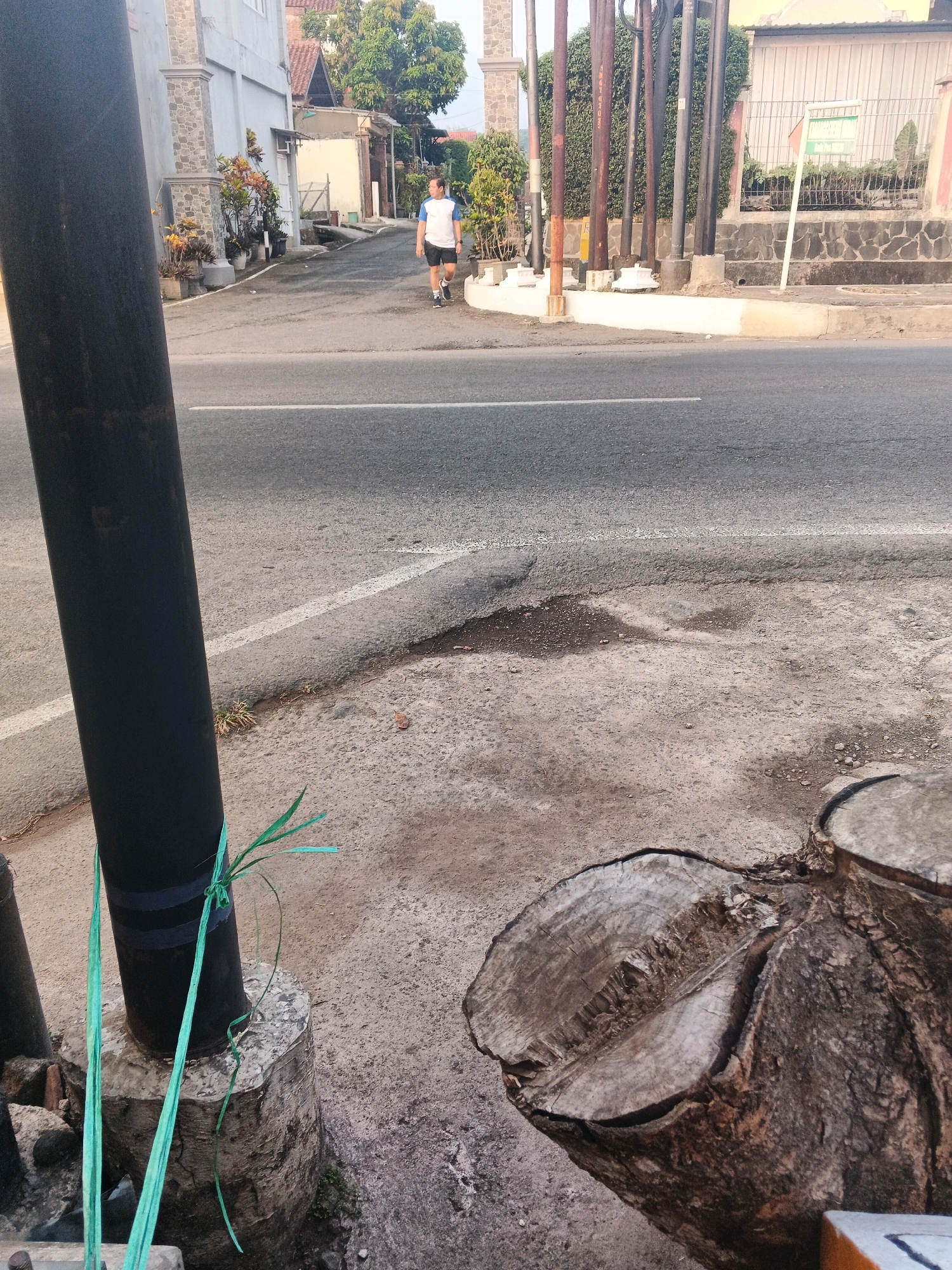 A man is walking on the sidewalk next to a street in Semarang, Indonesia. The man is wearing a white t-shirt with blue trim and black shorts. He is walking away from the camera. In the foreground, there is a large tree stump and a black pole wrapped with green tie-downs. The street is paved and has a white line down the center. In the background, there are buildings and trees. The sky is overcast.