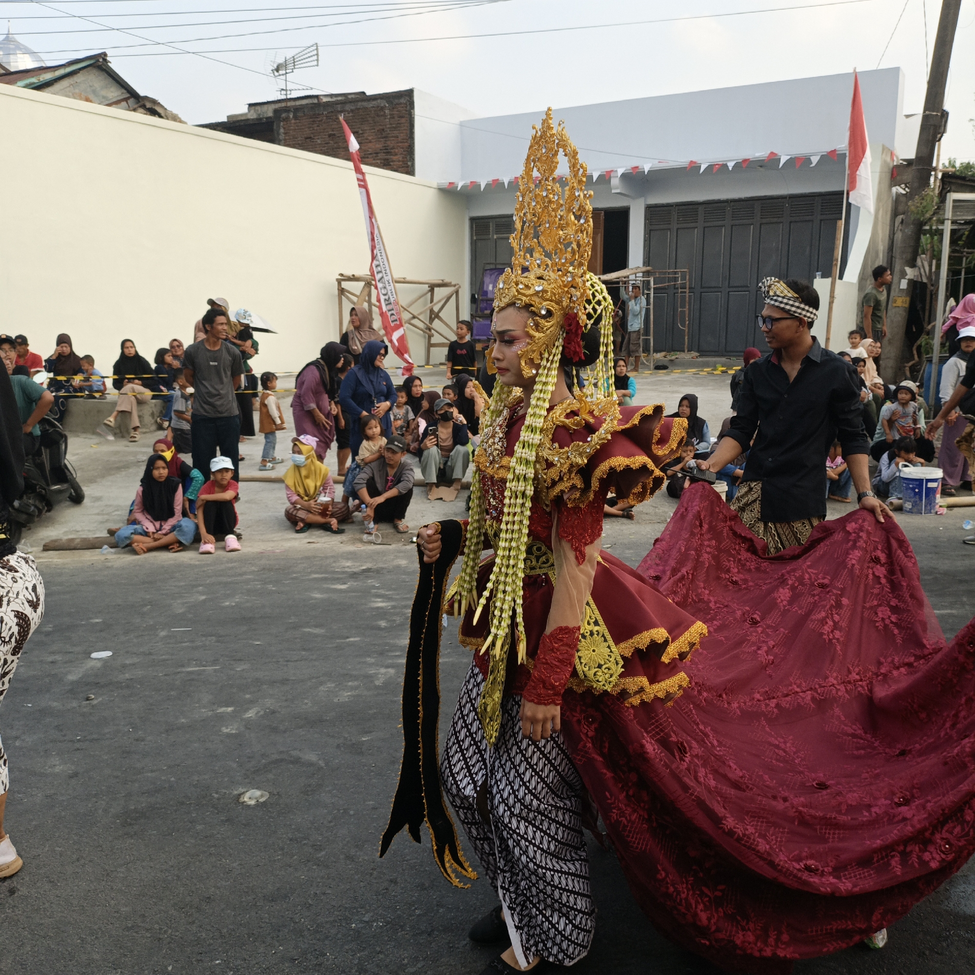 In Randudongkal, Indonesia, a cultural parade is underway on a street. The foreground is dominated by two individuals. A person in the center wears an elaborate, gold headdress adorned with intricate details and beads, a red and gold ornate costume with long, flowing sleeves, and a patterned lower garment. They are holding a dark, textured fabric. To their right, another person in a black shirt and patterned waist cloth stands holding a large, deep red, embroidered fabric, possibly part of the same costume or a prop.
The background is filled with spectators, seated and standing along the sides of the street, observing the parade. The buildings in the background are simple structures with white and light blue facades, some decorated with red and white triangular flags, indicating a national celebration. A red and white Indonesian flag is prominently displayed on a wooden structure. The overall environment suggests a public event taking place in a modest urban or semi-urban setting. The ground is paved asphalt.