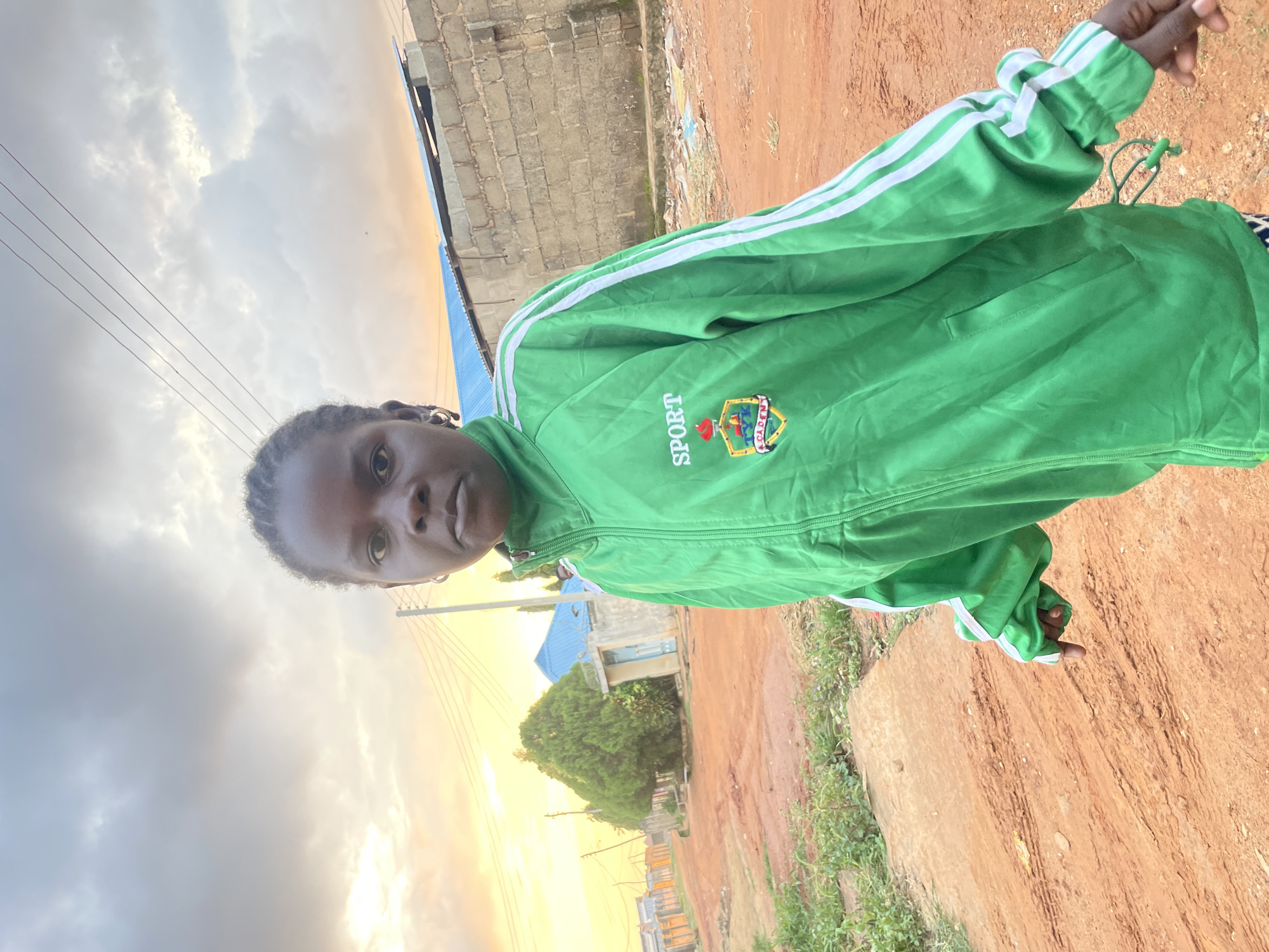A young girl stands in the foreground, facing the camera. She has dark skin and her hair is styled in braids. She is wearing a green jacket with white stripes on the sleeves and a crest on the left chest that reads "SPORT ACADEMY". The background is a street scene in Rido, Nigeria, with red dirt roads, a few buildings, and trees. The sky is cloudy with a hint of sunset light. The girl is pointing to her left, towards the viewer's left. The overall lighting suggests it is late afternoon.
