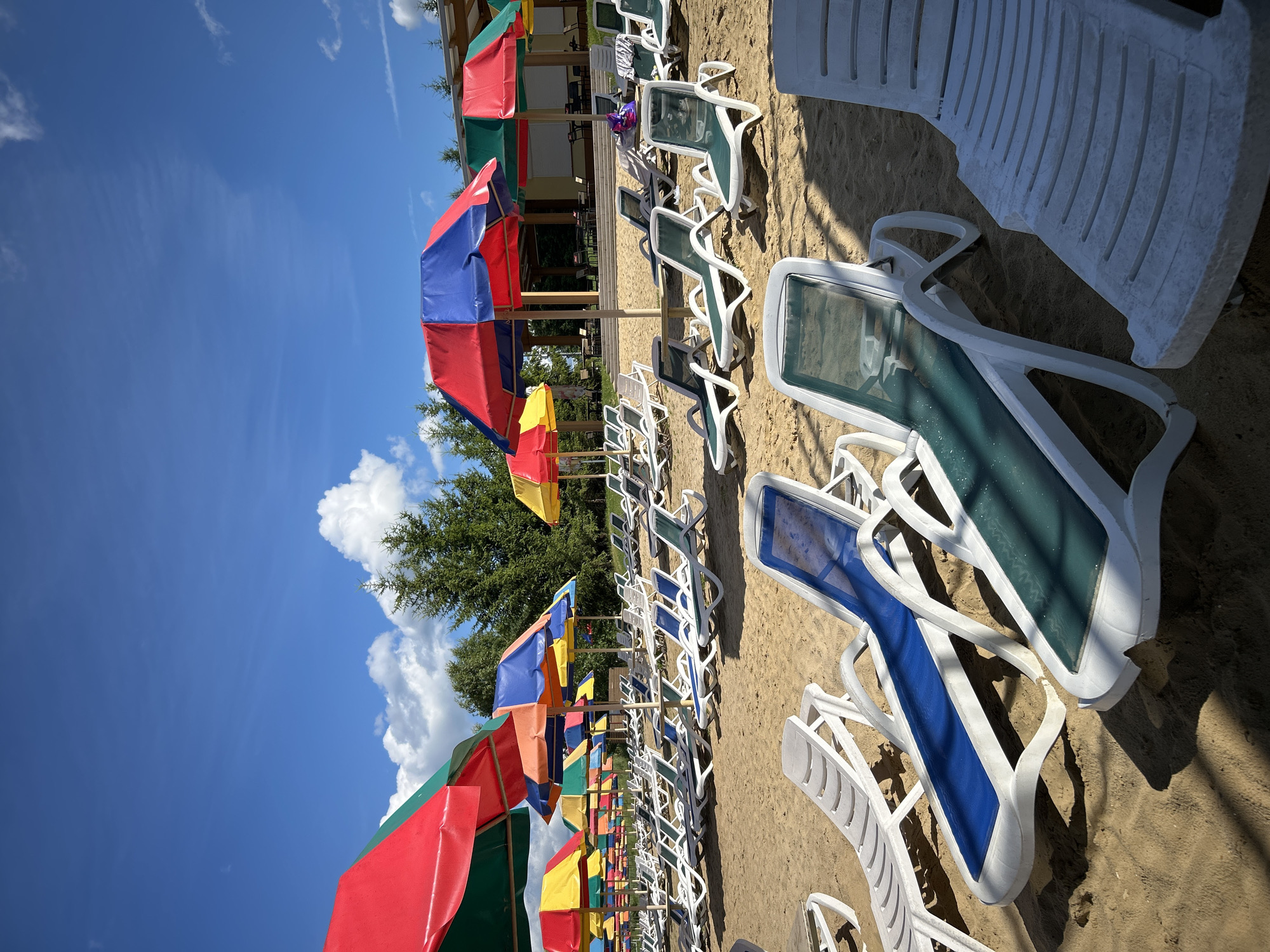 The image depicts a sandy beach with numerous empty lounge chairs arranged in rows under colorful umbrellas. The umbrellas are a mix of red and blue, red and green, and yellow and red, providing shade on a sunny day. The beach is backed by a line of trees and a building with a covered outdoor seating area. The sky is clear blue with a few scattered white clouds. The overall impression is one of a recreational area, likely a public beach or resort, on a pleasant day. It is not possible to determine the specific location within Yubileynyy, Russia, based on the visual information provided.