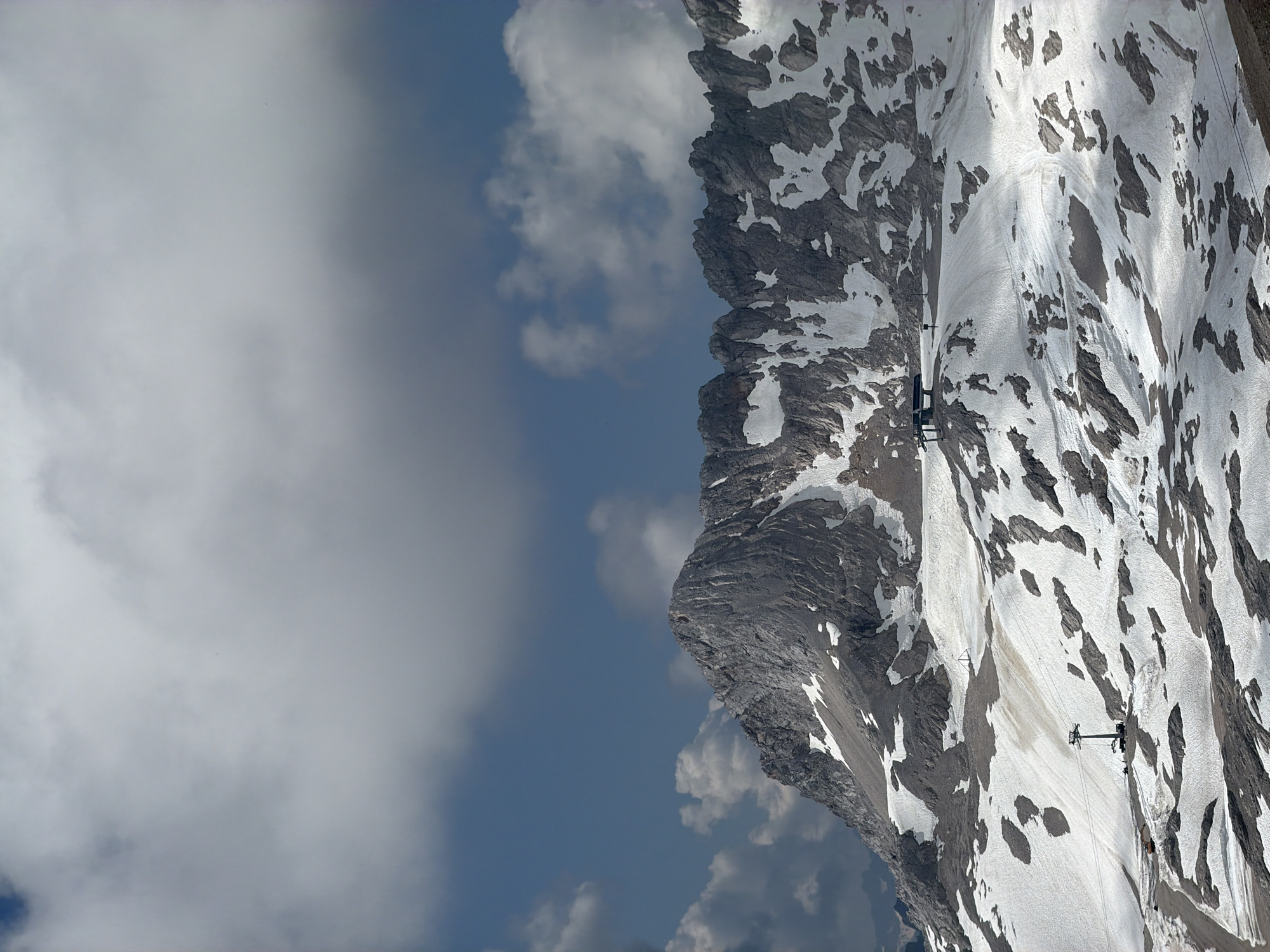 This image depicts a mountainous landscape with snow patches and rugged rock formations under a partly cloudy sky. In the foreground, there is a ski lift tower and station situated on a snowy slope. The area appears to be a ski resort or a similar alpine recreational area. The scale and features suggest a high-altitude environment. The image does not contain any people or any specific landmarks that would allow for precise identification of the location within Garmisch-Partenkirchen, Germany. The overall impression is one of a natural, high-altitude setting with infrastructure for tourism.