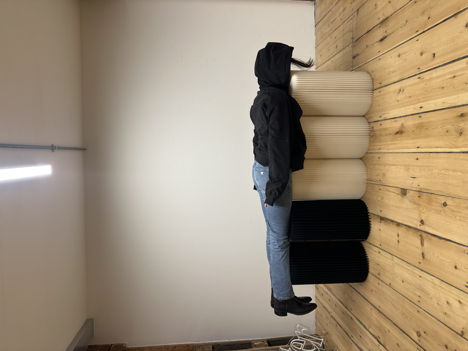 The image displays an indoor scene, likely a room or studio, with a person standing beside a stack of textured objects. The perspective is from the side, capturing the person's left profile.
On the left side of the frame, there is a light-colored, possibly off-white or beige, painted wall. A horizontal, bright white light fixture, appearing to be a fluorescent tube or LED strip, is mounted on this wall. Above the light, a silver-colored pipe or conduit runs horizontally along the wall. The wall extends into a corner on the right.
The right side of the frame features a wall constructed from light-colored wooden planks, arranged horizontally. The floor is also made of similar wooden planks, but they are oriented perpendicularly to the wall planks.
A person is standing upright, facing towards the left side of the frame. They are wearing a black hooded sweatshirt with the hood pulled up, partially obscuring their face. A strand of dark hair is visible peeking out from under the back of the hood. They are dressed in light blue denim jeans, and the word "GUESS" is distinctly visible as a patch or stitching on their right back pocket. On their feet, they are wearing black ankle boots.
To the right of the person, stacked against the wooden wall, are five distinct, oblong, ribbed objects. The top three objects are a cream or light beige color, while the bottom two are black. These objects appear to be soft or pliable, possibly functioning as modular seating or decorative partitions. They are stacked vertically, reaching approximately to the height of the person's shoulders or head.
On the wooden floor in the bottom right corner, a few miscellaneous items are visible, including what appear to be tangled white objects, possibly hangers or wires, and some small debris. The overall lighting suggests an indoor setting, likely illuminated by artificial light from the fixture on the wall.