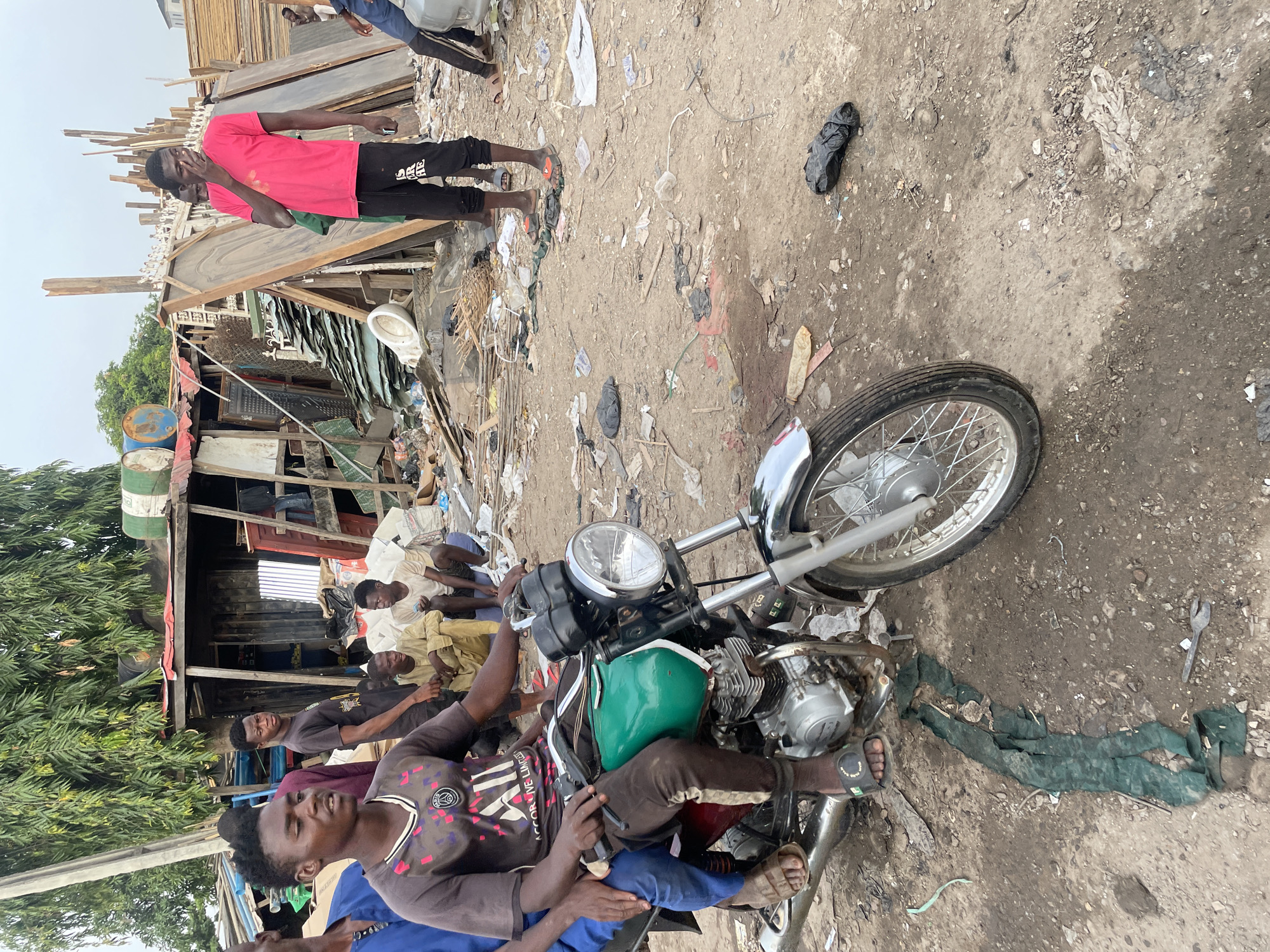 This image captures a vibrant, outdoor scene in what appears to be an informal settlement or market area in Abuja, Nigeria, under a bright, possibly overcast daytime sky.
In the foreground, a silver and green motorcycle is a central subject, with a person sitting on it. This individual wears a dark, patterned t-shirt that features text like "PSG" (likely referring to Paris Saint-Germain), "CORRUPTION LIMITED", and "ARE YOU READY". Another person sits closely behind them on the motorcycle. The ground is unpaved dirt, heavily covered with a variety of litter including plastic bags (one black, one resembling a discarded condom), paper, fabric scraps, and other debris. A black slipper and a metal wrench are also visible on the ground.
Several individuals, mostly men, are present in the scene. To the mid-left, a group of men are seated on makeshift benches or on the ground, some wearing light-colored clothing. Further back on the right, a man in a bright pink t-shirt and black sweatpants, with the letters "ARE" visible on his leg, stands and appears to be touching his face.
The background is dominated by various makeshift structures constructed from corrugated metal sheets, wooden planks, and other salvaged materials. Blue and green barrels are visible near some of these structures, alongside piles of wooden doors and what appears to be a white ceramic toilet bowl. Green foliage from trees or bushes is visible on the far left, contrasting with the man-made structures and debris. The overall impression is one of a busy, active community living or working in a densely packed and resourcefully built environment.