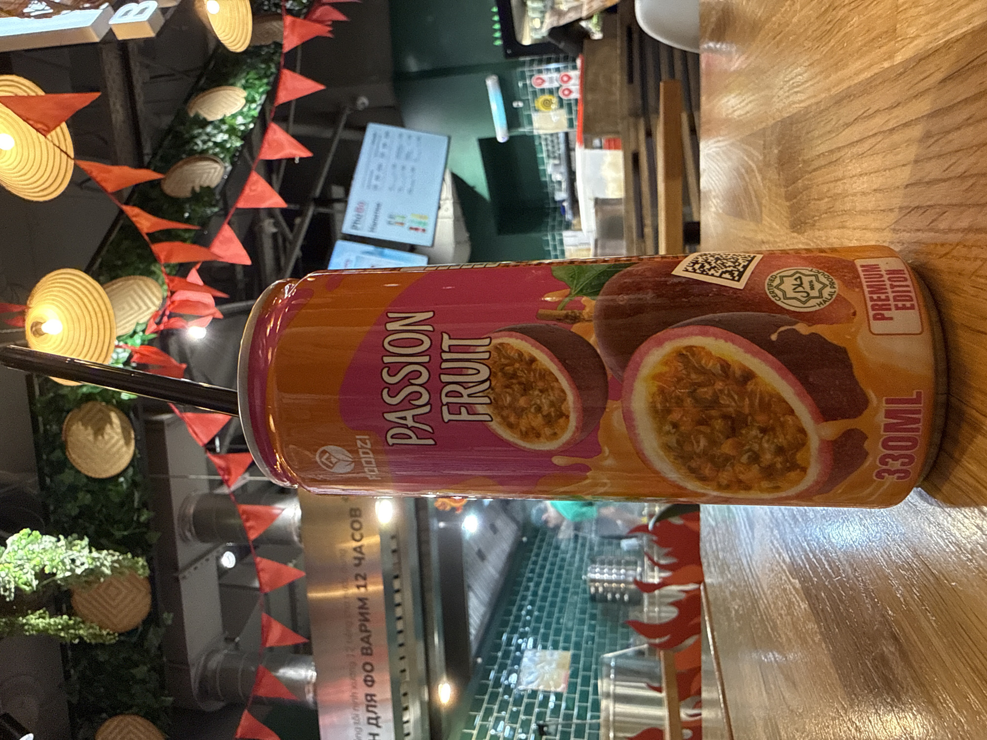 A beverage can with an orange and pink label featuring passion fruit imagery and text reading "FOODZI PASSION FRUIT 330ML PREMIUM EDITION" stands on a wooden table. A black straw is inserted into the top of the can. In the background, a restaurant interior is visible, with green tiled walls, stainless steel equipment, and a menu board displaying "PhoBo". Red triangular flags are strung across the upper part of the background. The location is identified as Rostov-on-Don, Russia.