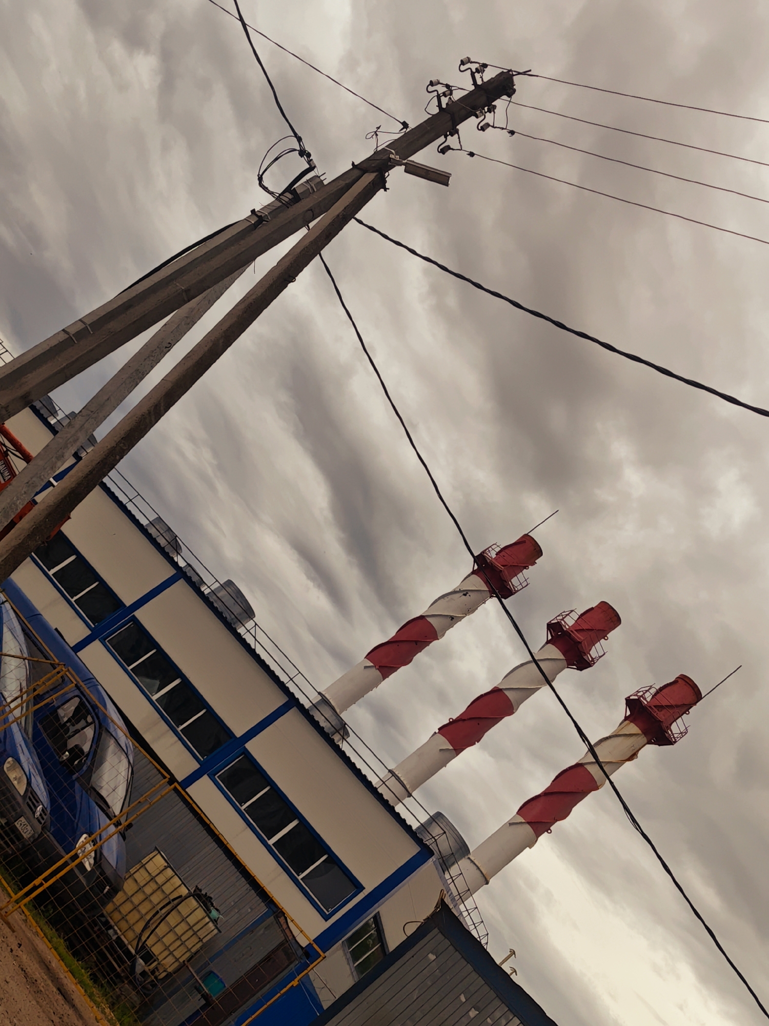 The image depicts an industrial scene in Zaprudnya, Russia, under a cloudy sky. A concrete utility pole with multiple wires stands prominently, angled against the sky. To the right, three large, white and red striped chimneys rise diagonally. Below the chimneys and the utility pole is a building with a white facade and a blue trim. The building has several rectangular windows with dark frames. A blue van is partially visible on the left side of the image, parked behind a yellow metal fence. The ground appears to be dirt and gravel. The overall atmosphere is industrial and somewhat bleak, likely due to the overcast weather.