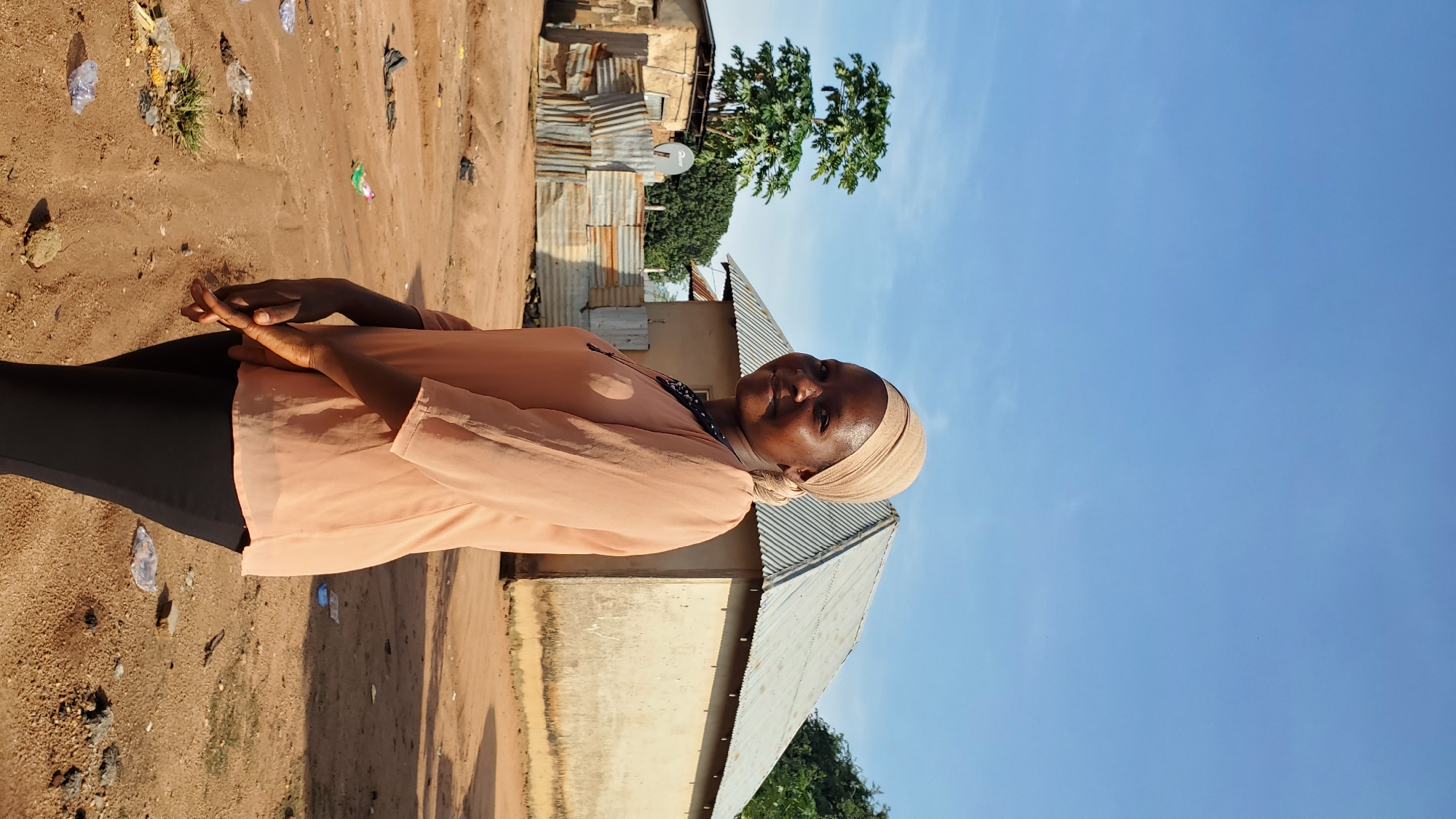 In Jalingo, Nigeria, a Black woman wearing a peach-colored headscarf and a matching short-sleeved blouse stands facing the camera. She is also wearing black pants. Her hands are clasped in front of her. Behind her, a small, light-colored building with a corrugated metal roof is visible. To the left of the building, there is another structure with a corrugated metal fence and a satellite dish. The ground is sandy and littered with debris. The sky is clear and blue.