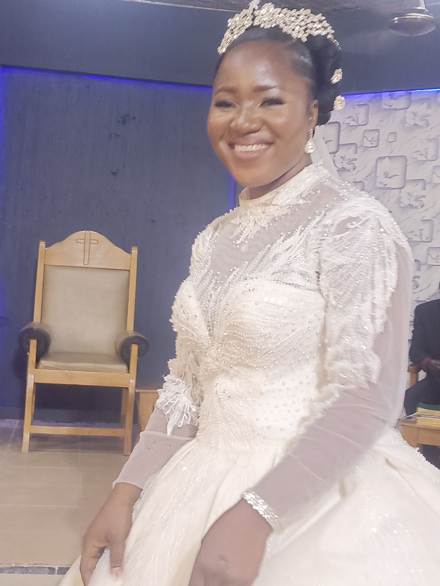 Akwanga, Nigeria: A smiling woman in a white, embellished wedding gown and tiara stands in an interior space. A wooden chair with a cross on the back is behind her. The walls are a combination of blue and a textured white. A man can be seen in the background, seated near a table. The floor appears to be tile.