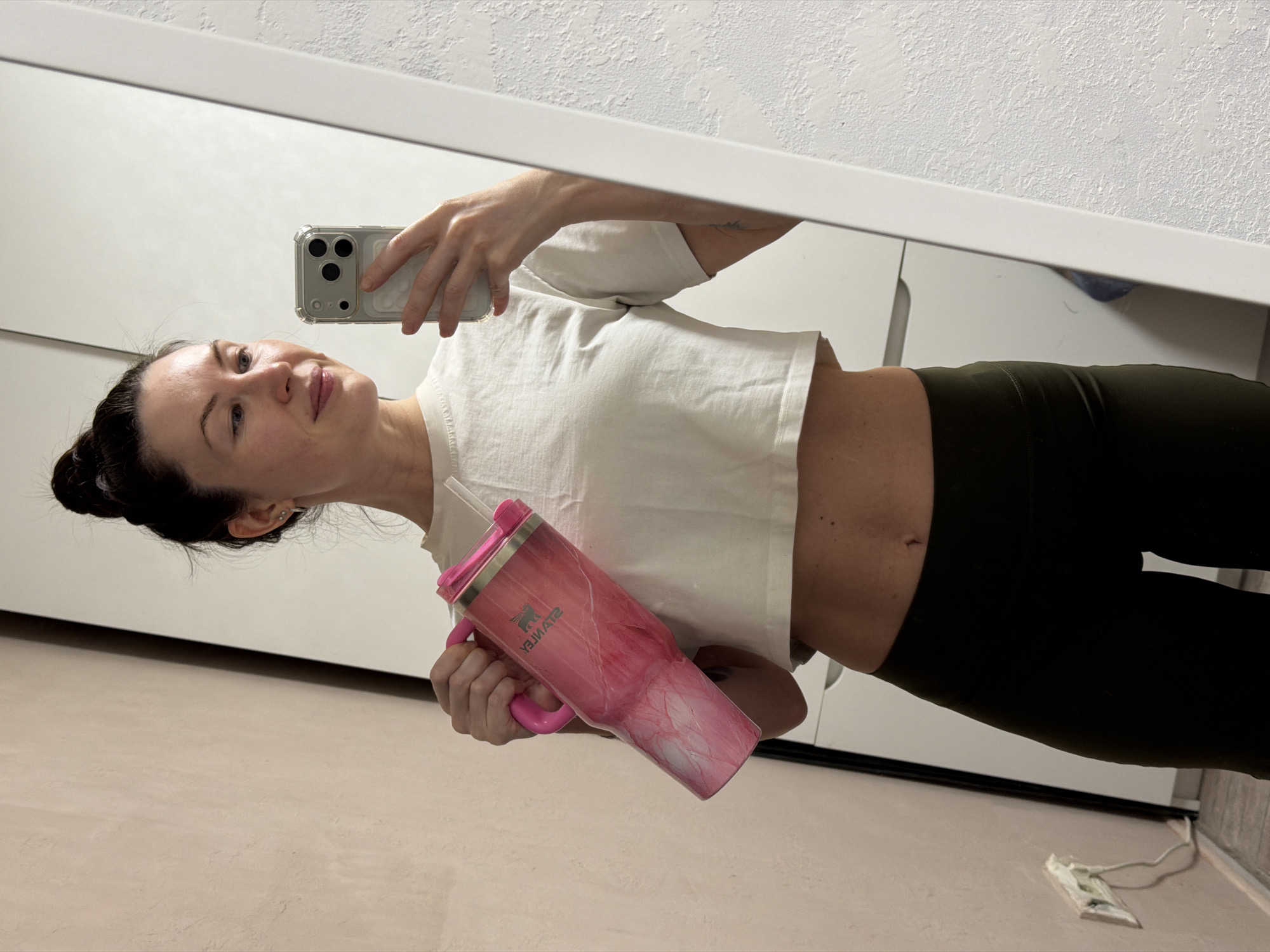 A person is taking a selfie in front of a mirror. The person is holding a pink water bottle in their left hand and a cell phone in their right hand. The person is wearing a white crop top and dark green leggings. The person has dark hair pulled back in a bun. The background appears to be a room with white walls and furniture. The person is located in Perm, Russia.