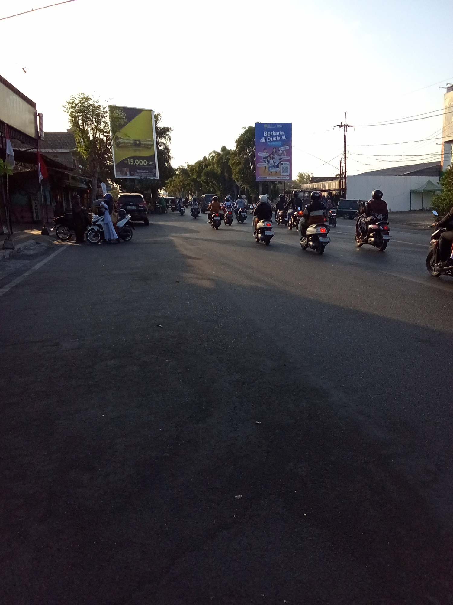 The image captures a street scene in the city of Kediri, Indonesia, during what appears to be late afternoon or early evening, given the soft, warm lighting and long shadows. The road is asphalted and shows signs of moderate traffic. A prominent feature of the scene is a large billboard on the left side of the road with a yellow and green design and the text "15.000-an". Further down the street, another billboard is visible, this one blue, displaying text in Indonesian that translates to "Work in the AI World," with images of people and a QR code.
The street is populated with numerous motorcycles, indicating a common mode of transportation in this area. Several individuals are riding these motorcycles, some with passengers. They are dressed in casual attire, and many are wearing helmets. On the left side of the road, there are buildings and shops, one of which has a red and white flag displayed. The presence of trees and utility poles suggests an urban environment. The overall impression is a bustling street life typical of a city in Indonesia.