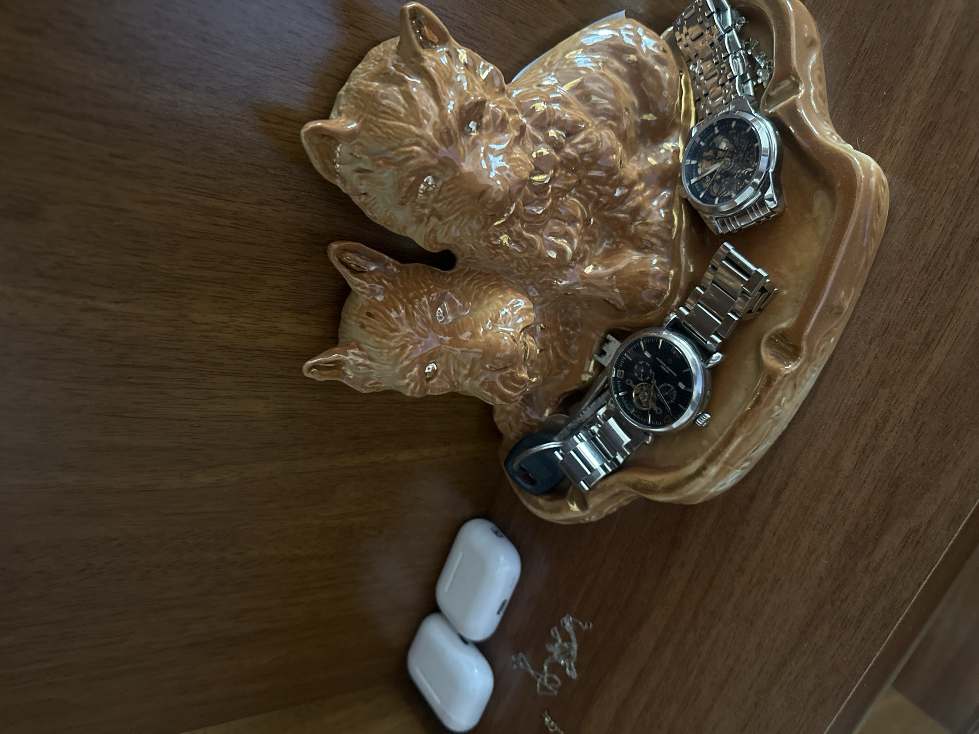 The image presents a top-down view of a wooden surface, on which rests a ceramic dish in the shape of two intertwined fox figures. The dish has a glazed, golden-brown finish. Inside the dish, there are two wristwatches with stainless steel bands, both having dark-colored faces. Adjacent to the dish, outside its rim, are a pair of white wireless earbuds, along with a fine silver chain. The location of the scene is unspecified (Unknown, Unknown).