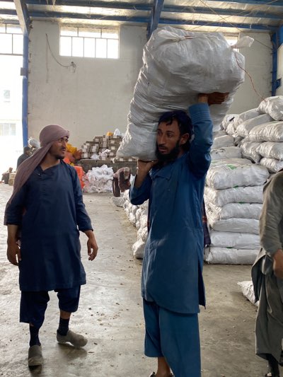 Inside a large warehouse in Shurandam, Afghanistan, two men are visible. One man, wearing a blue tunic and pants, carries a large white sack over his shoulder. The second man, also dressed in blue, is slightly to the left, his head covered with a tan cloth. The warehouse contains numerous stacks of white sacks or bags, suggesting storage or processing. Boxes are also stacked in the background. The scene appears to be a workspace, with a focus on manual labor involving the transportation of large bags.