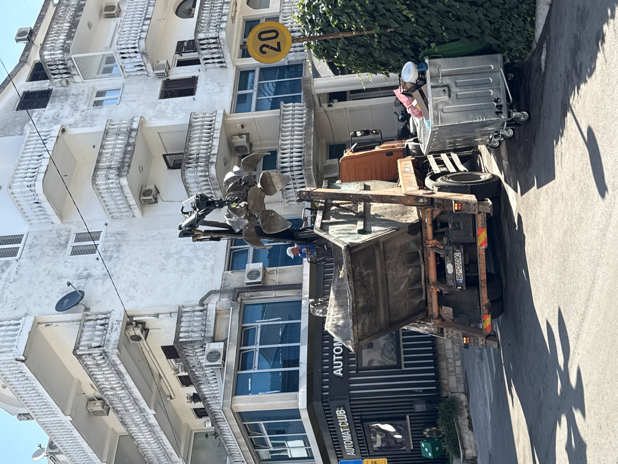 An industrial garbage truck with a claw mechanism is parked on a street in Budva, Montenegro, next to a building with multiple balconies. The truck is actively collecting waste from a large dumpster. A person wearing a white cap and blue shirt is visible in the elevated cabin of the truck, operating the claw. Adjacent to the dumpster, there is a large, silver wheeled bin filled with trash bags and various discarded items. A yellow circular sign with the number "20" is mounted on a pole to the right of the scene. The building appears to be a multi-story residential or commercial property. The street surface is asphalt, and there is a distinct shadow cast on it from an unknown object. The area is well-lit by daylight.
