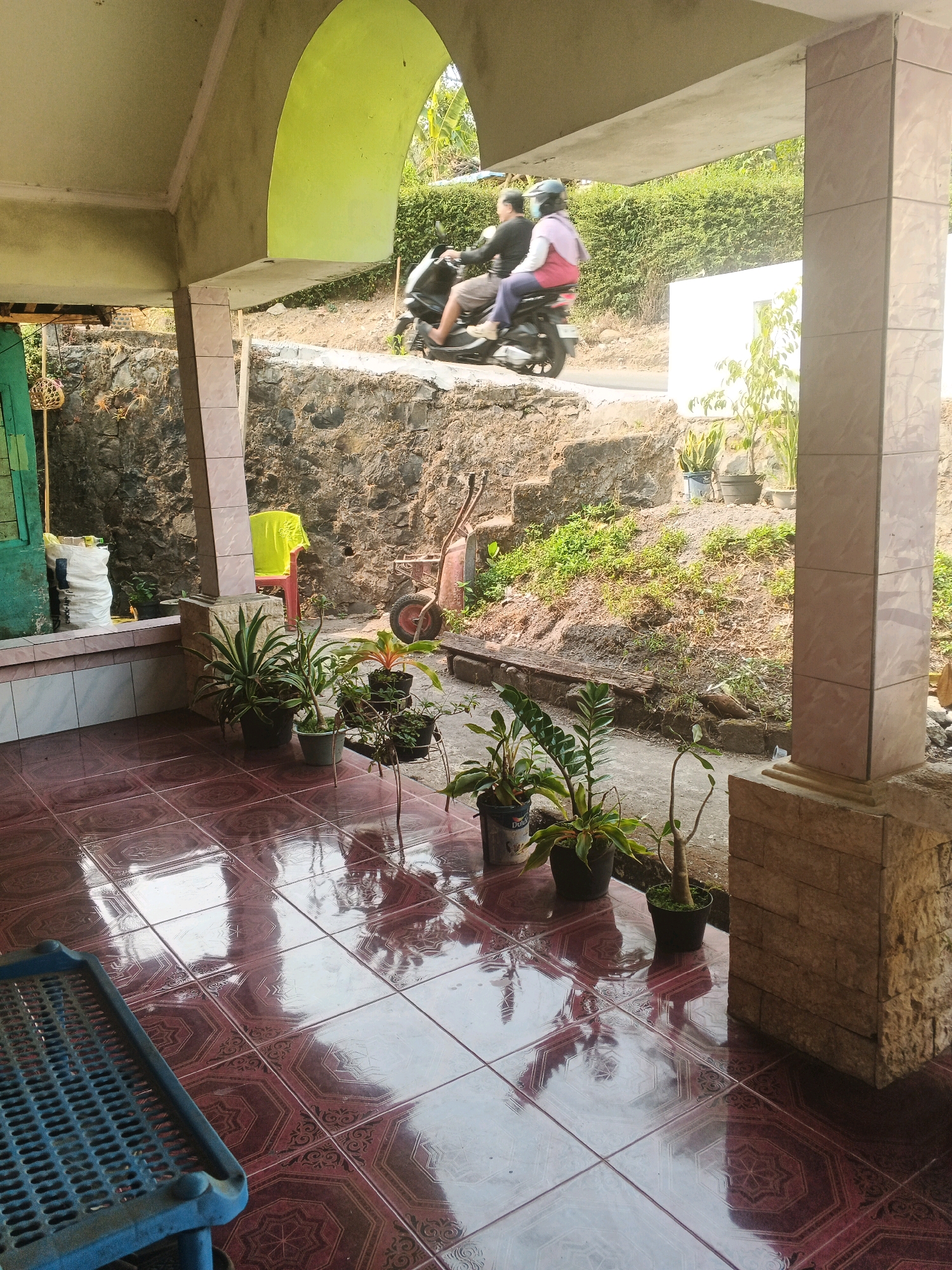 This image depicts a scene on a porch in Keji, Indonesia. Two individuals, a man and a woman, are riding a motorcycle on a paved road in the background. The woman is wearing a helmet and a pink top, while the man is wearing a dark shirt. The porch itself has tiled flooring with a reddish-brown pattern. Several potted plants are arranged on the porch. To the left, a blue plastic object with a grid pattern is visible. In the background, there is a stone wall, a green structure, a red chair with a yellow shirt draped over it, and a wheelbarrow. The environment suggests a rural or semi-rural setting with vegetation and a hillside.