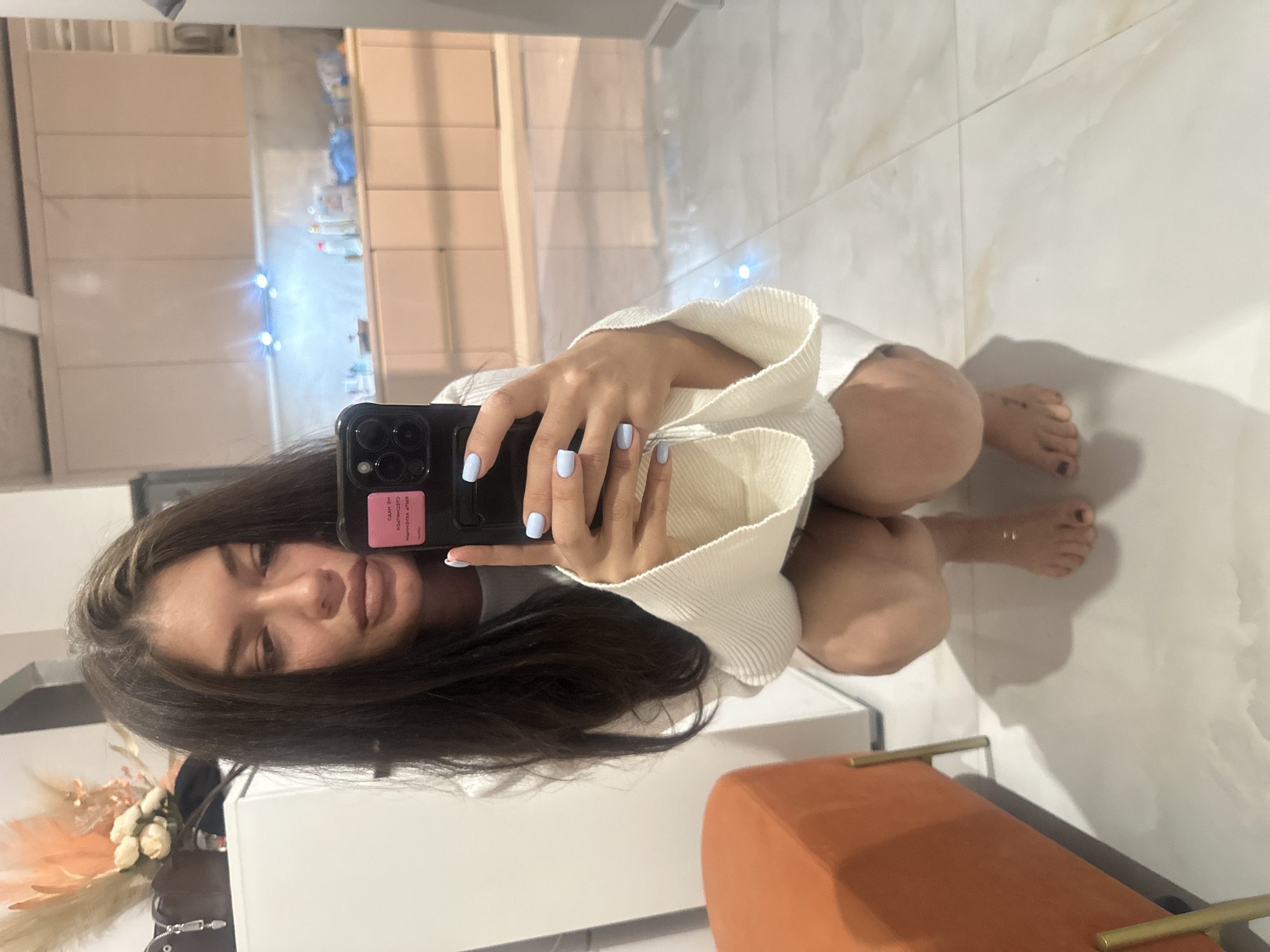 A young woman is posing for a selfie in what appears to be a bathroom. The selfie is taken in a mirror. She is wearing a white, ribbed sweater and has long, dark hair. Her fingernails are painted light blue. She is holding a black smartphone with a pink sticker on the back. The bathroom has light-colored tile floors and white cabinets. A floral arrangement sits on the cabinet surface. In the mirror's reflection, one can see the bathroom's contents and possibly some decorations. There is an orange upholstered bench with gold-colored legs/handles at the bottom of the picture. The location, Khimki, Russia, cannot be determined from the information available in the image.