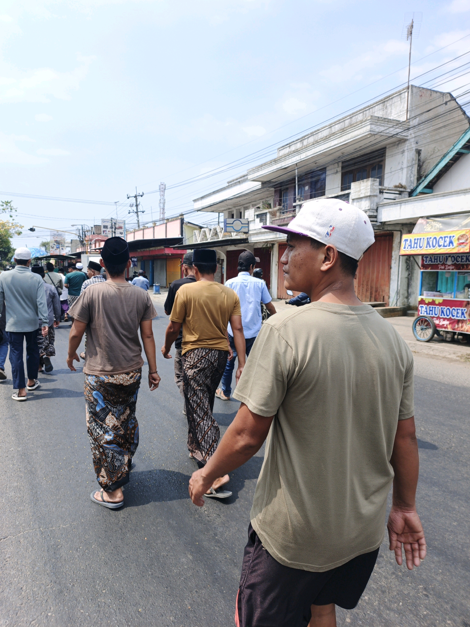 A group of men walk down a paved road in Mojoduwur, Indonesia. The man in the foreground, facing away from the camera, wears a light green t-shirt and black shorts. He has a light-colored baseball cap with a purple brim on his head. Behind him, several other men are also walking away from the camera. Some are wearing traditional Indonesian sarongs and peci caps. One man in the middle ground wears a brown t-shirt and a patterned sarong. Another man to his right wears a golden-brown t-shirt and a patterned sarong. On the right side of the road, a food cart labeled "TAHU KOCEK" is visible, with a blue-wheeled cart attached. Buildings line the left side of the road, some appearing to be shops with closed metal shutters. The sky is clear and blue with scattered white clouds. Power lines and utility poles are visible above the buildings.