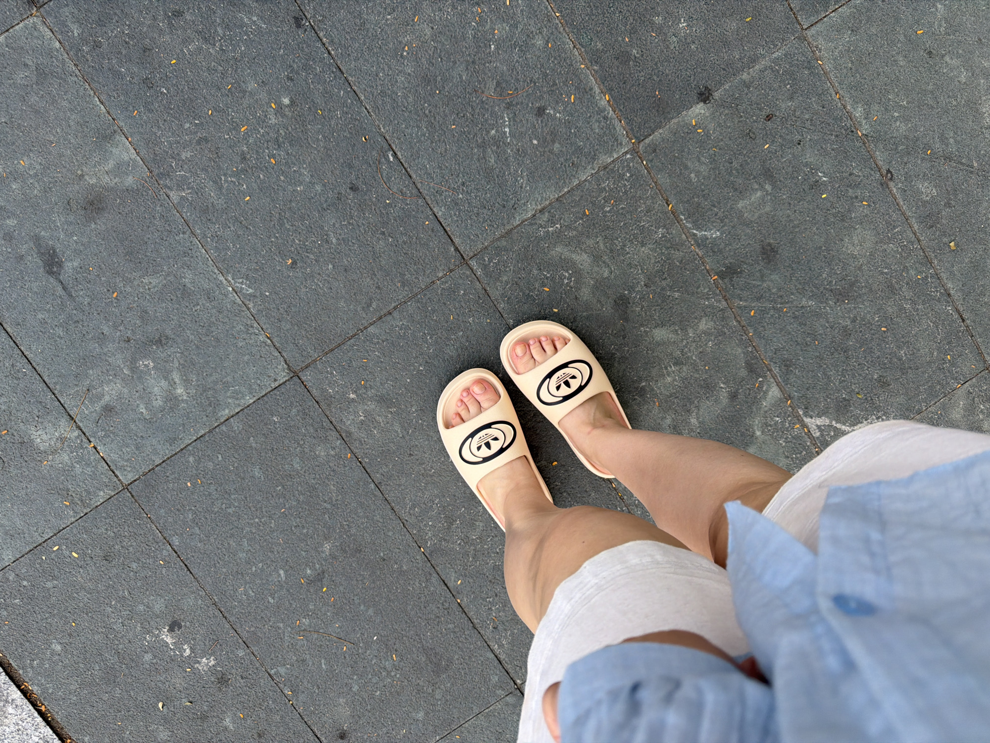 A top-down, eye-level shot shows a person standing on a paved surface, possibly in Nha Trang Ward, Vietnam. The paved surface consists of dark gray rectangular stones arranged in a grid pattern, with a few scattered, small yellow objects. The person is wearing light beige slides with a black logo on top, and their feet are visible. They are also wearing white shorts and a light blue shirt is partially visible. The focus is on the person's feet and the surrounding paved surface.