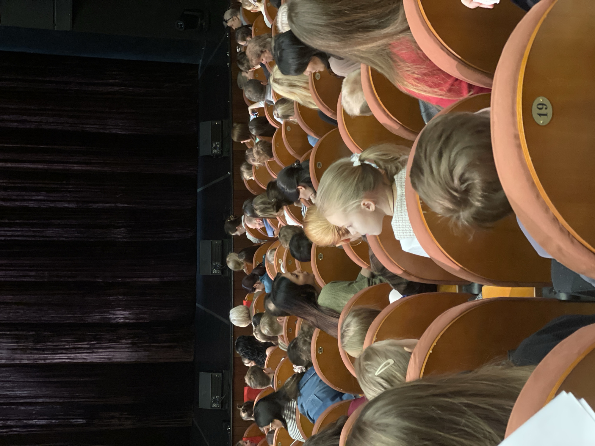 The image shows an audience seated in rows of upholstered chairs, possibly in a theater or auditorium. The chairs are curved, and some have seat numbers visible. The audience consists of people with various hair colors and styles, most facing forward, presumably watching a performance. A dark curtain or backdrop is visible to the left, and some electronic equipment is visible on the wall between it and the chairs. The location is Unknown, Unknown.