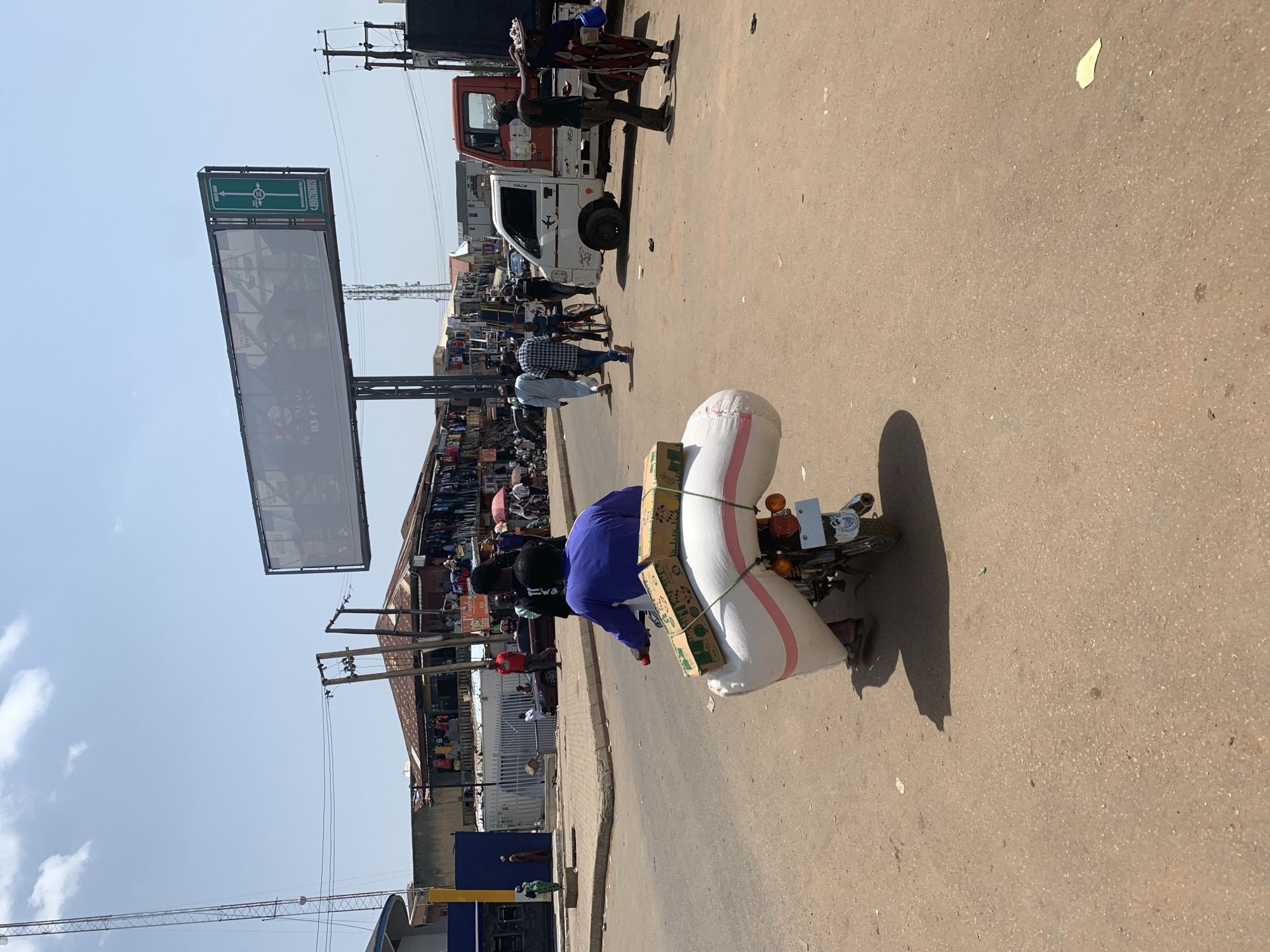 A wide shot captures a sunny day in Jos, Nigeria, likely in a busy market area. In the foreground, a person wearing a blue shirt rides a motorcycle laden with two large, white, cylindrical objects tied down with rope, along with two cardboard boxes. The motorcycle has a license plate. The road surface is a light brown, dusty asphalt.
In the midground, several people are walking. To the left, a man in a blue and white patterned shirt walks with a bicycle. Further back, a small white truck with a red cab is parked on the right side of the road. A person stands beside the truck, holding a tray of items. In the background, a strip of shops with awnings is visible, displaying clothing and other goods. A large billboard on a metal frame stretches across the upper midground, displaying an advertisement, and to its right, a green directional sign for "BYPASS ROAD." A telecommunications tower stands tall on the left side of the image, and telephone poles with wires are visible throughout the scene. The sky is clear blue with a few scattered white clouds. The overall impression is of a bustling urban environment.
