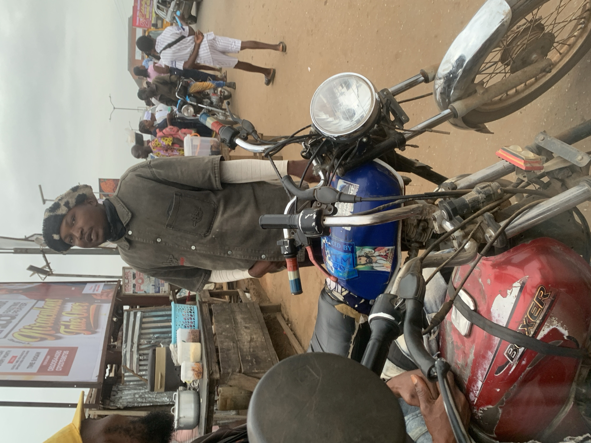 In Shagamu, Nigeria, a man wearing a patterned beret and a grey button-up shirt stands next to a motorcycle. The man appears to be speaking. Behind him, several other people are visible, along with stalls selling various items, including food and drinks. The scene is outdoors, with a dusty, unpaved surface and a cloudy sky overhead. The motorcycle in the foreground is red and blue, with a sticker of Jesus on the fuel tank. The overall atmosphere suggests a busy market or roadside scene.