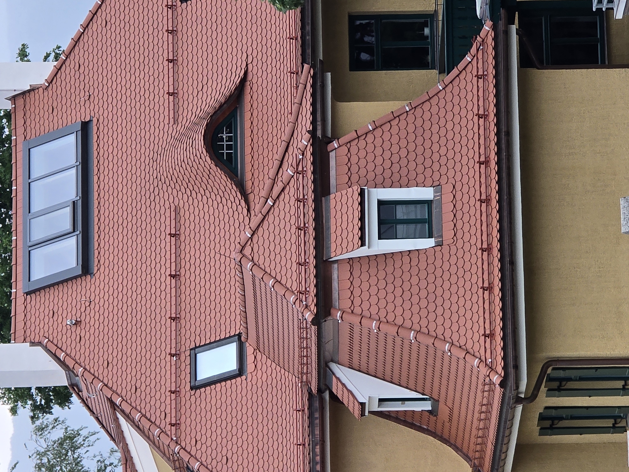 The image shows a close-up view of a red tiled roof with several dormer windows and skylights. The roof is made of overlapping scale-shaped tiles. In the center of the roof, there is a distinctive triangular dormer window with dark green trim and a decorative grille. To the left and right of this central dormer are other dormer windows with white trim and dark green windows. Two rectangular skylights are visible on the upper part of the roof, set within a dark grey frame. A white chimney rises from the left side of the roof. The building appears to be a residential property with a yellow facade visible below the roofline. The overall impression is of a well-maintained, traditional building.