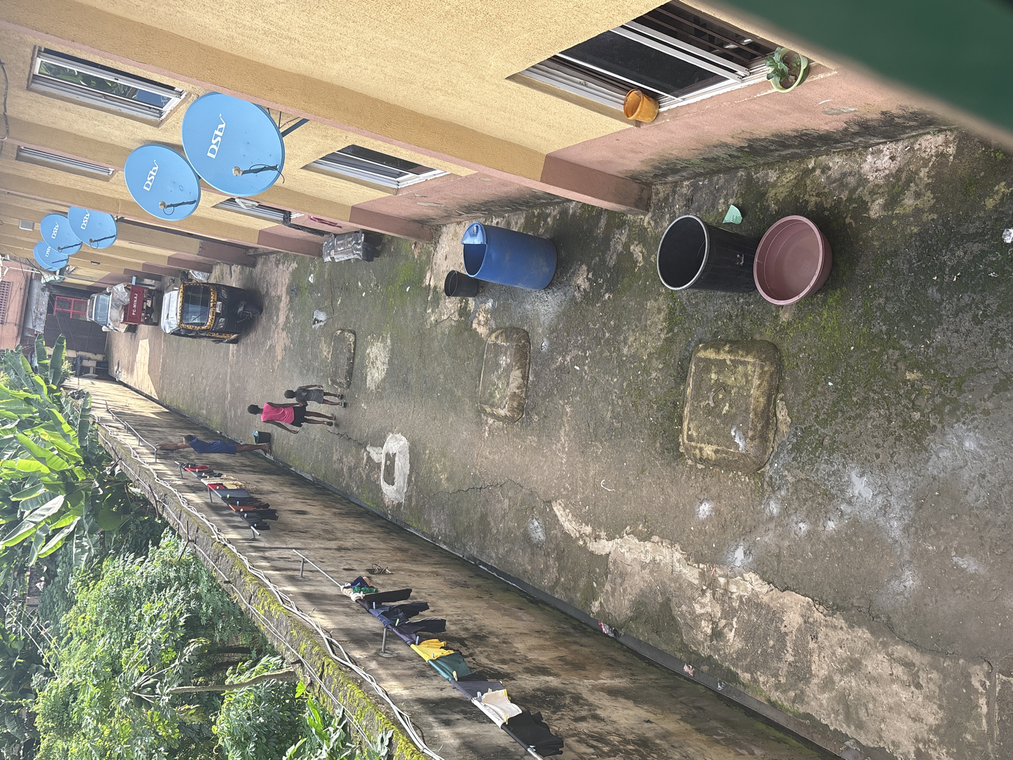 The image depicts an alleyway or courtyard located in Onitsha, Nigeria, likely in a residential or commercial area. On the left, a concrete wall runs along the length of the scene, with laundry hung on a clothesline suspended from it. Several items of clothing, including shirts and pants, are visible. Lush green vegetation, including banana trees, grows beyond the wall on the left.
In the alleyway, two children, a girl in a pink top and shorts and a younger child, walk away from the camera. A man in a blue shirt and dark shorts stands near the wall, appearing to be attending to the laundry. Further down the alley, a three-wheeled auto-rickshaw (a Keke Napep) is parked. Across the alley, a yellow and brown multi-story building with a pink and yellow facade is visible. Several blue satellite dishes, marked with "DSTV," are mounted on the building's exterior. Windows with white frames are also present.
On the ground of the alley, there are several large blue and black plastic barrels, and a brown plastic basin. The ground is paved with concrete and appears damp and mossy in places, with some debris scattered about. In the distance, another vehicle, possibly a white pickup truck with a red stripe and "PC-MINAJ" written on it, is parked. A small potted plant sits on a windowsill of the building. The overall impression is one of a busy, lived-in urban environment.