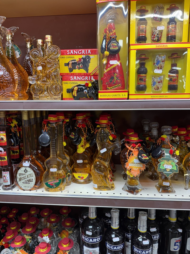 The image shows shelves stocked with bottles of Spanish liquor in what appears to be a shop in Fuentes de Oñoro, Spain. The shelves display uniquely shaped glass bottles, including designs resembling guitars, horses, and traditional Spanish figures. Labels on the bottles indicate that some contain "Moscatel" and "Sangria." The bottles are adorned with miniature red hats, and some are packaged in sets with matching shot glasses. The presence of "Bodegas Garces, S.A." on packaging points to one of the manufacturers. In the bottom row, there are multiple bottles of "Ricard." Price tags are visible on many of the items. The shelving is made of a light brown wood.