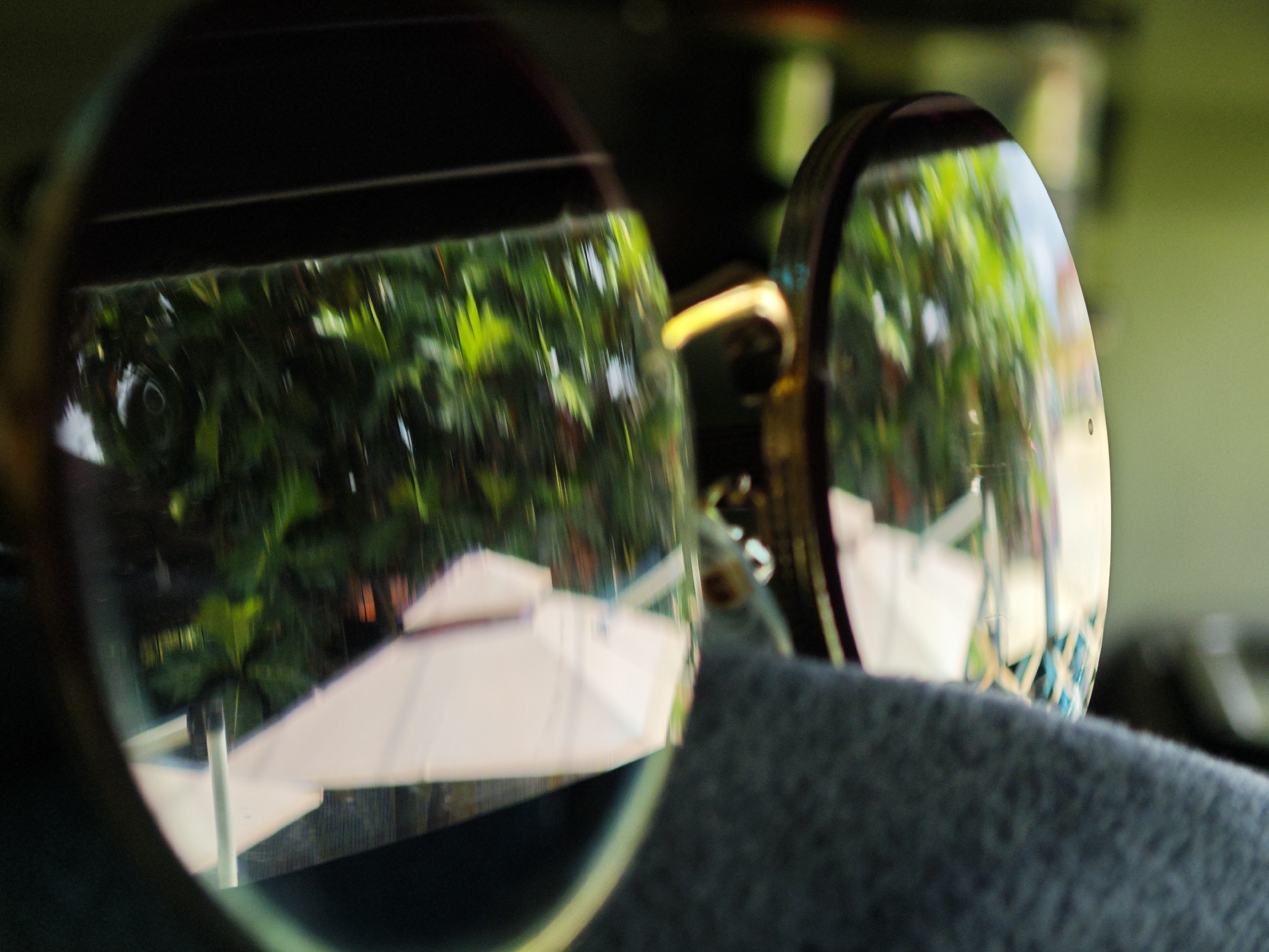 A close-up, shallow depth-of-field photograph captures the reflection of an outdoor scene in two lenses of a pair of sunglasses. The sunglasses are resting on a dark gray, textured surface, possibly fabric. In the left lens, the reflection shows lush green foliage and what appears to be the edge of a white umbrella structure. The right lens reflects a similar outdoor environment, with more pronounced green leaves and a lighter, brighter area that could be a building or sky. The framing suggests the sunglasses are being worn or held by someone indoors, looking out at a bright, leafy environment, possibly a resort or garden area in Bờ Y, Vietnam. The overall impression is one of a bright, sunny day viewed through tinted lenses.