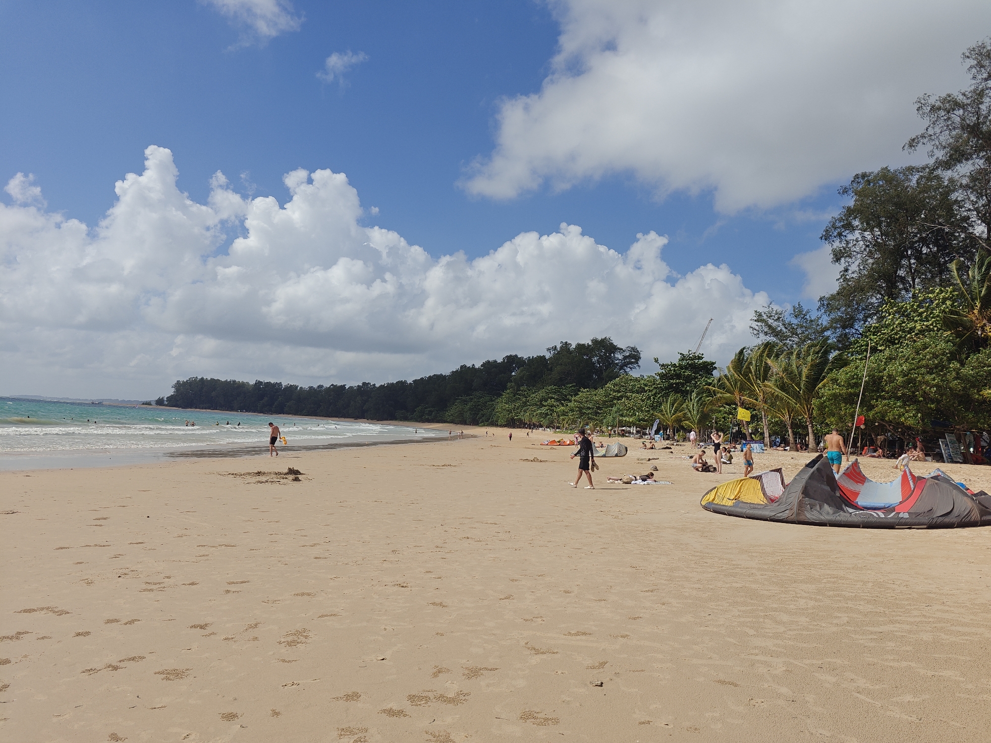 The image depicts a wide, sandy beach scene in Sakhu, Thailand, under a bright blue sky with scattered white clouds. The ocean is visible in the distance, with gentle waves rolling onto the shore. A line of lush green trees and palm trees forms the backdrop along the coastline.
Several people are present on the beach. In the foreground, a large, colorful kite is laid out on the sand. To its right, a man wearing blue shorts is standing. Further down the beach, closer to the water, a person in black shorts is walking, and another person is lying on the sand. In the mid-ground, closer to the water, a few individuals are standing and wading in the shallow ocean. Further down the beach, groups of people are scattered, some relaxing on the sand and others near tents and what appear to be other kitesurfing equipment. The sand itself is marked with numerous small mounds, likely created by sand crabs.