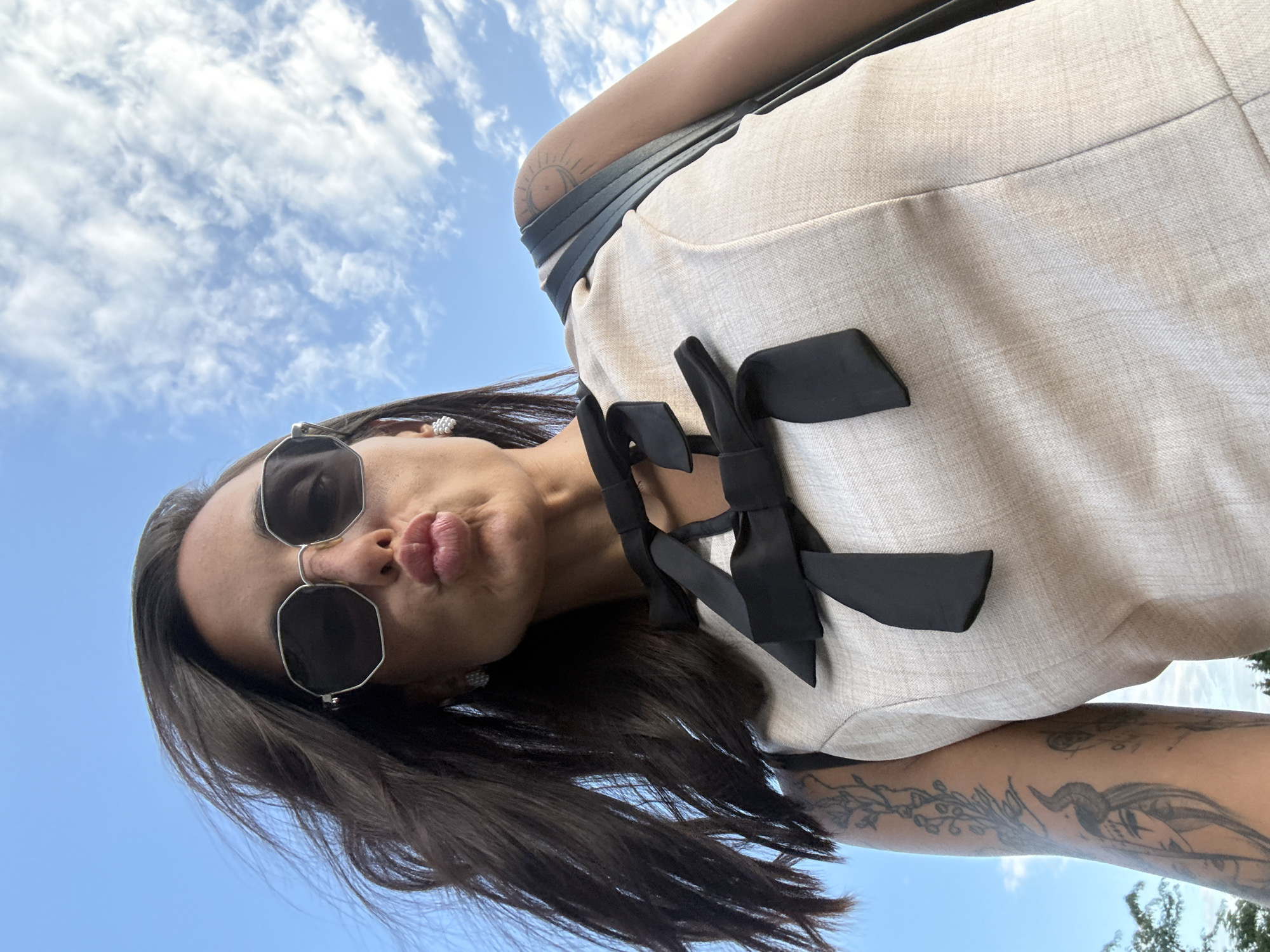 The image shows a low-angle, close-up selfie of a woman outdoors with a blue sky and scattered clouds in the background. She is wearing sunglasses, a light-colored dress with black bow detailing, and a black bag strap is visible over her shoulder. She has dark hair and visible tattoos on her arm. Her lips are pursed, as if blowing a kiss. The image was taken in Pleven, Bulgaria.