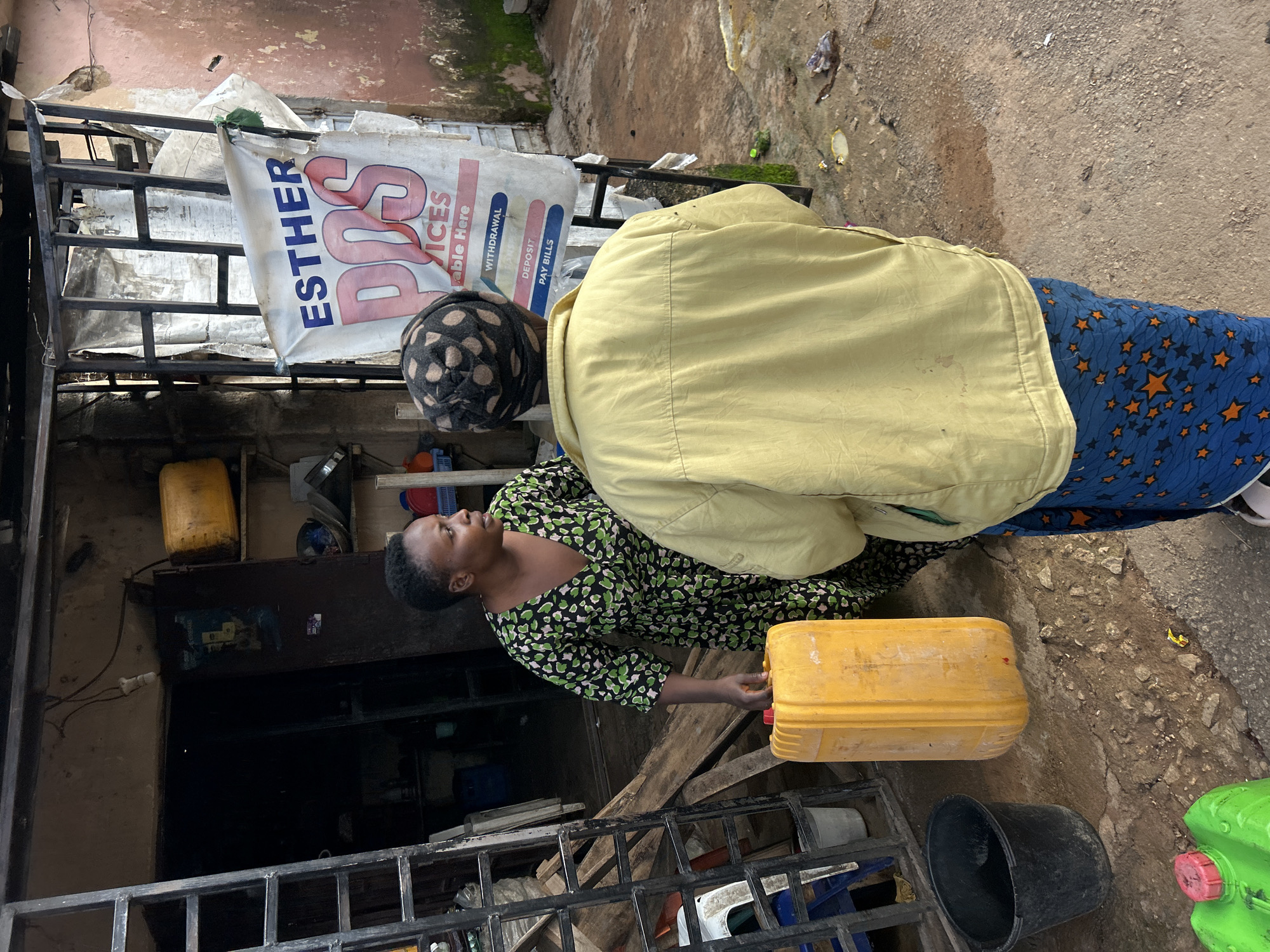 In Jos, Nigeria, outside a modest shop with a metal gate and a sign that reads "ESTHER POS SERVICES", a woman in a green and black patterned dress is handing a large yellow jerrycan to another person. The second person, wearing a yellow jacket and a patterned headscarf, has their back to the camera. The ground is dirt and scattered debris. Other objects visible include a black bucket, a green jerrycan, and shelves filled with items inside the shop.