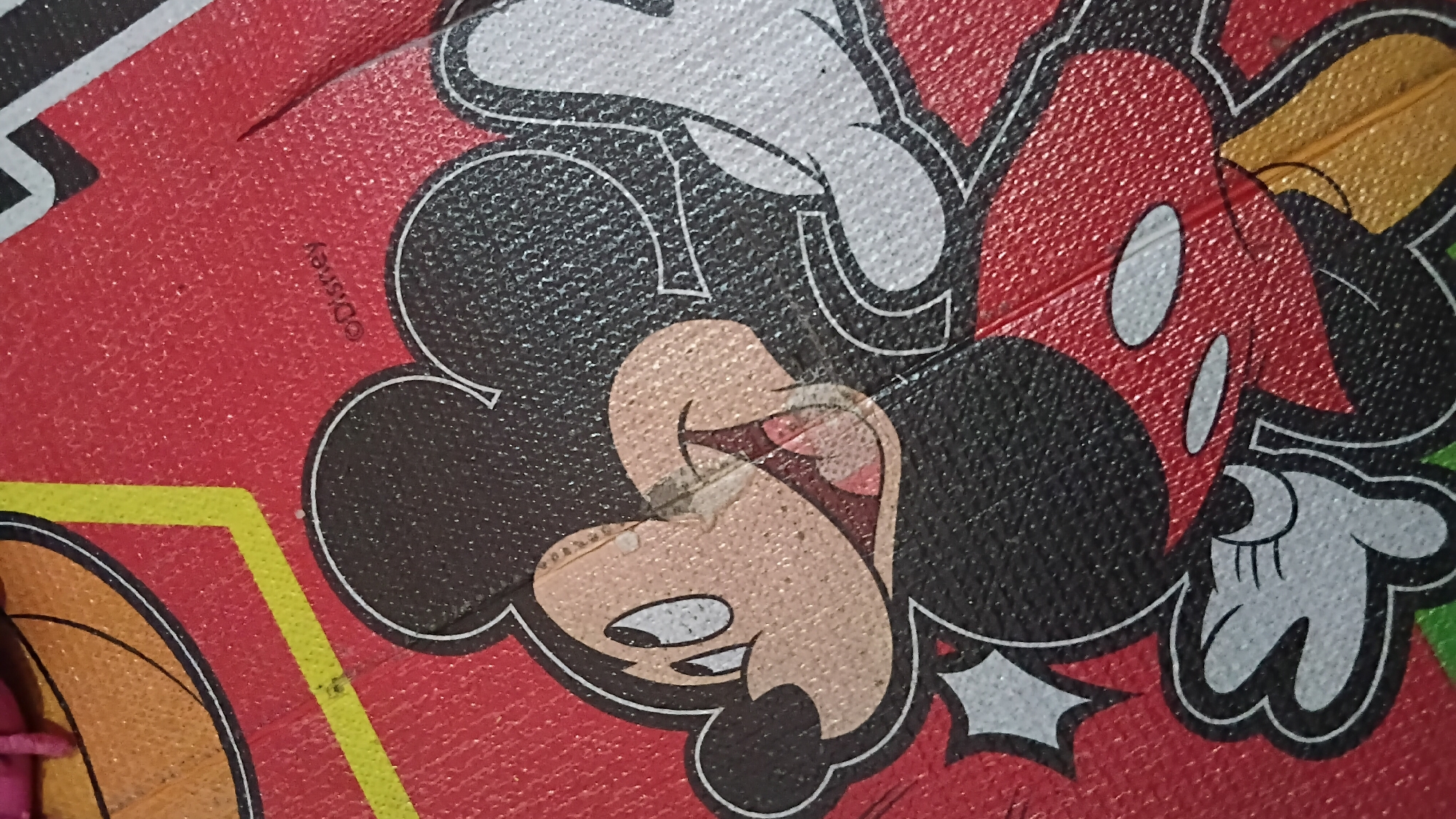 The image shows a close-up view of a red surface with a depiction of Mickey Mouse. The texture of the surface appears to be slightly rough or pebbled. Mickey Mouse is shown in his classic red shorts with white buttons, black ears, and white gloves. His face is light brown with large, expressive eyes and a wide, open smile showing teeth. There is a slight scuff or tear on his cheek. The background is a solid red color with a subtle texture. In the top left of the image, a portion of a yellow and black graphic element is visible, possibly part of a larger design or border. Near the top right of the image, the word "Disney" is visible as a watermark or copyright notice, printed in small, white, stylized letters. The image is not taken in the city of Prabumulih, Indonesia, or any other specific city. It appears to be a close-up of a product or artwork featuring the Disney character.