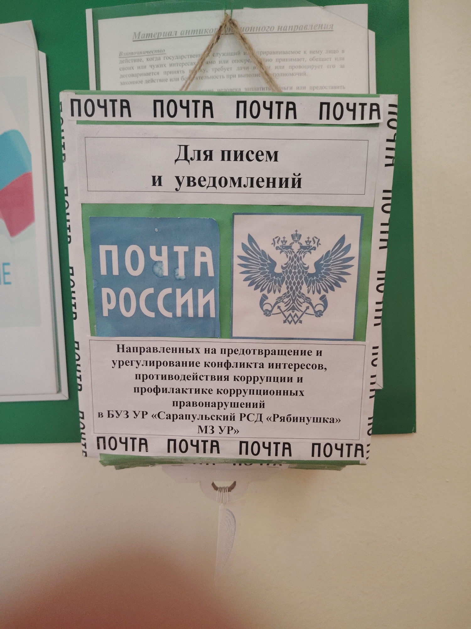 The image displays an indoor wall display, likely in a public or institutional setting within Sarapul, Russia. The primary subjects are an informational poster and an attached suggestion box, designed to resemble a mailbox.
The top portion of the display board is titled "Материал антикоррупционного направления" (Anti-corruption material). Below this title, there's a text defining "Взяточничество" (Bribery), describing it as an act by a public servant or similar individual who, in their own or others' interests, directly or indirectly accepts, promises, or demands a bribe for performing or omitting duties.
Attached below this poster is a pocket-like box, labeled repeatedly with the word "ПОЧТА" (Mail). Its main heading states "Для писем и уведомлений" (For letters and notifications). This box features two distinct emblems: a blue square with "ПОЧТА РОССИИ" (Russian Post) in white, and to its right, a blue and white depiction of the Russian double-headed eagle.
The explicit purpose of this "mailbox" is detailed in the text below the emblems: "Направленных на предотвращение и урегулирование конфликта интересов, противодействия коррупции и профилактике коррупционных правонарушений в БУЗ УР «Сарапульский РСД «Рябинушка» МЗ УР»" (Aimed at preventing and resolving conflicts of interest, countering corruption, and preventing corruption offenses in BUZ UR "Sarapulsky RSD 'Ryabinushka' MZ UR"). This indicates the box is specifically for submitting communications related to anti-corruption efforts within the mentioned institution. The lighting suggests it is daytime.