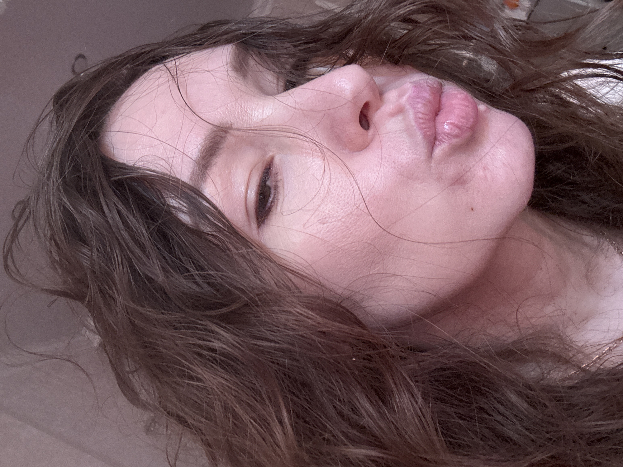 A close-up portrait, oriented on its side, features the right profile of a person's face, likely female, captured from a low angle looking upwards. The subject's hair, a rich dark brown, appears slightly damp or tousled, with individual strands falling across her forehead and cheek. Her right eye is partially open, looking towards the viewer. Her nose is prominent in profile, and her lips are pursed into a distinct pout, showing natural creases and a glossy sheen. The skin has a fair complexion with visible texture and a subtle pinkish tone. A faint metallic chain, possibly a necklace, is discernible near her neck on the right side. The background is an undifferentiated, muted brownish-grey, suggesting an indoor setting. The image evokes a casual and somewhat playful or contemplative mood. This portrait is set in Beryozovy, Russia, though no specific location cues are visible in the generic background. No text is present in the image.
