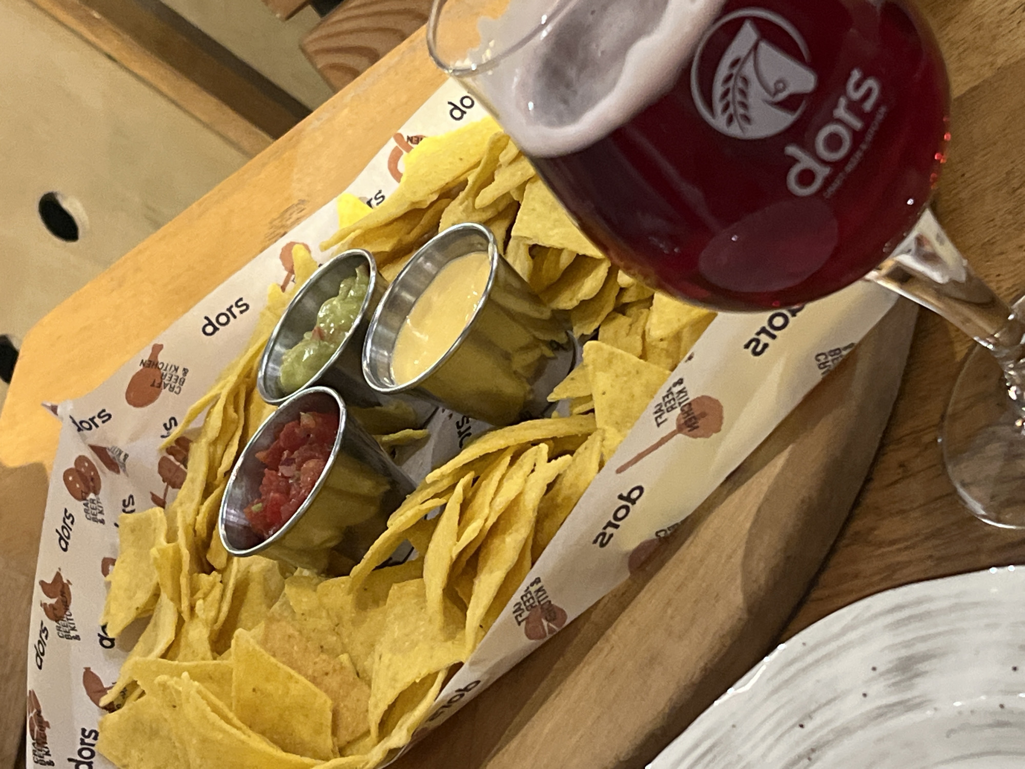 The image shows a plate of nachos with three different dips (salsa, guacamole, and cheese) along with a glass of beer. The branding on the beer glass and the printed paper lining the plate identifies the location as "dors Craft Beer & Kitchen." It is most likely located in Yerevan, Armenia, based on the background of the image.