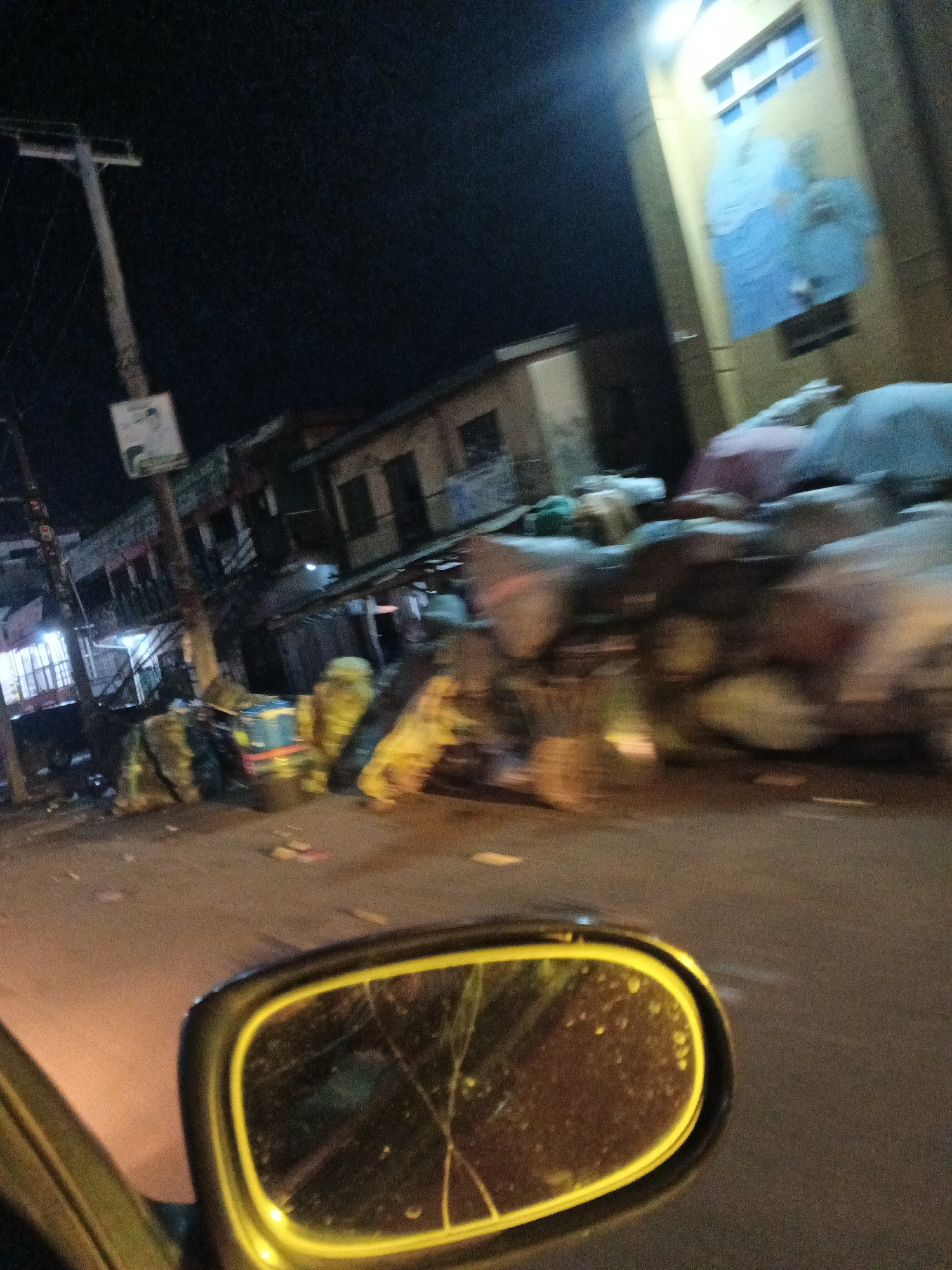The image depicts a street scene at night in Jos, Nigeria. A blurred view from inside a vehicle shows a side-view mirror in the foreground, reflecting some of the surroundings and the night sky. The street itself appears to be lined with buildings and what looks like market stalls or refuse piles, heavily blurred due to motion. A utility pole with a sign is visible on the left side of the street. The lighting suggests artificial illumination from streetlights or building lights. The overall impression is one of a busy, urban environment at night, possibly a market area or commercial street, with a sense of movement and a slightly gritty atmosphere.