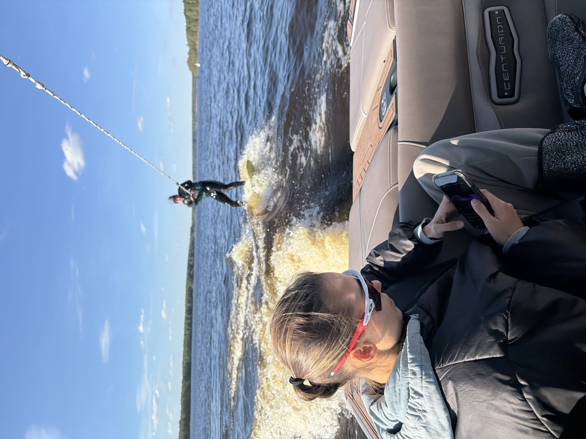 A sideways shot captures a scene of waterskiing from the perspective of someone on a boat, possibly the Centurion brand as suggested by the interior upholstery. A woman in a jacket is seen looking at her cell phone. In the background, a person is suspended in the air, attached to the boat by a long white tow rope, as they waterski across the water. The setting appears to be a lake or wide river with trees lining the distant shoreline, under a partly cloudy sky. The location of Gorodishche, Russia, is not visually discernable.