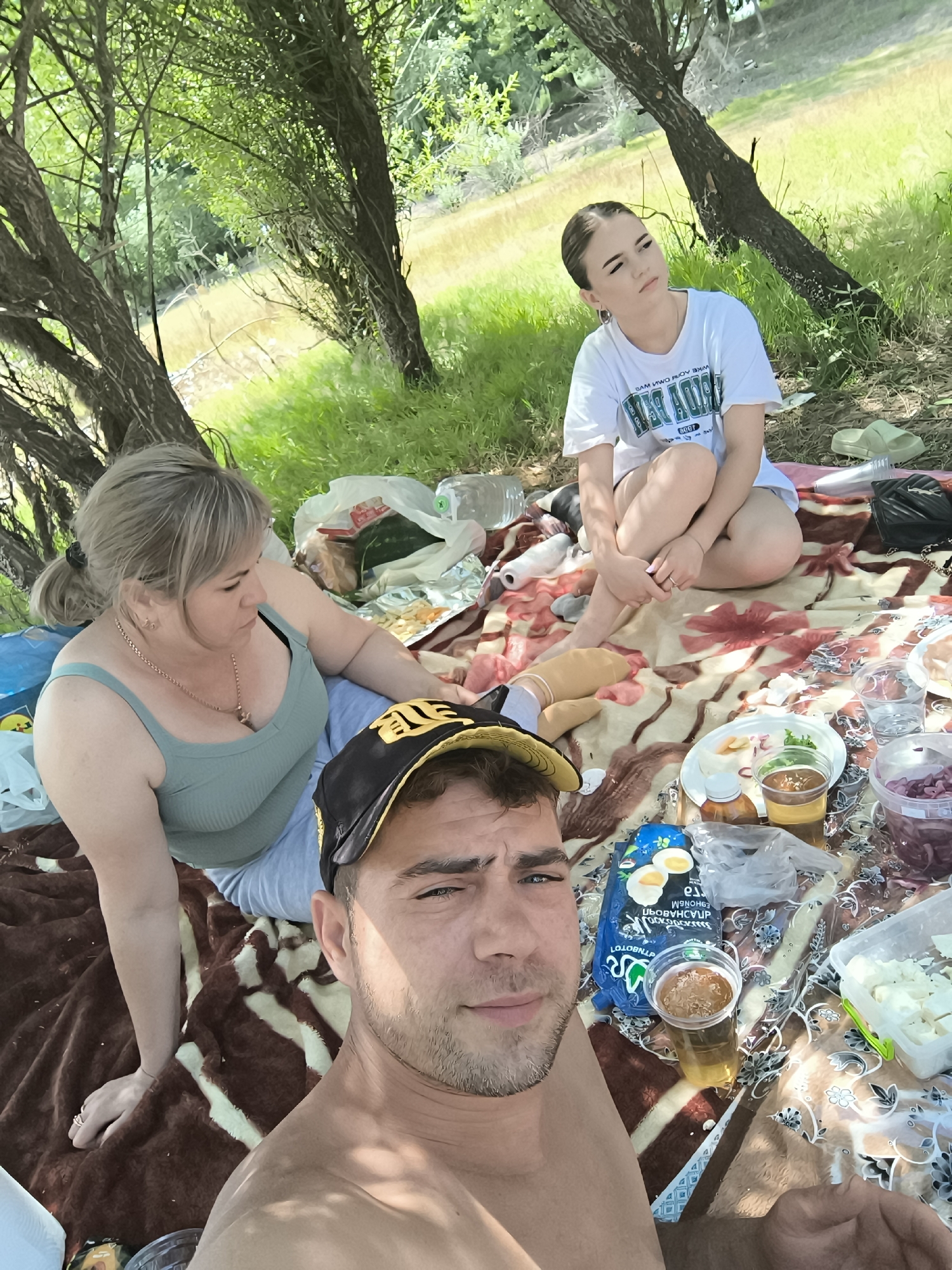 In Costești, Moldova, a group of three individuals are enjoying a picnic on a blanket spread on the ground, under the shade of trees. A man in the foreground, wearing a black baseball cap with yellow accents, faces the camera, his upper body visible. To his left, a woman with blonde hair pulled back in a ponytail, wearing a mint-green tank top and light blue pants, is seated, looking away from the camera. Further in the background, a young woman with dark hair styled up, wearing a white t-shirt and dark shorts, is seated with her legs crossed, looking upwards. The picnic spread includes various food items, drinks in plastic cups, and bags. The environment is natural, with lush green grass and trees surrounding the area.