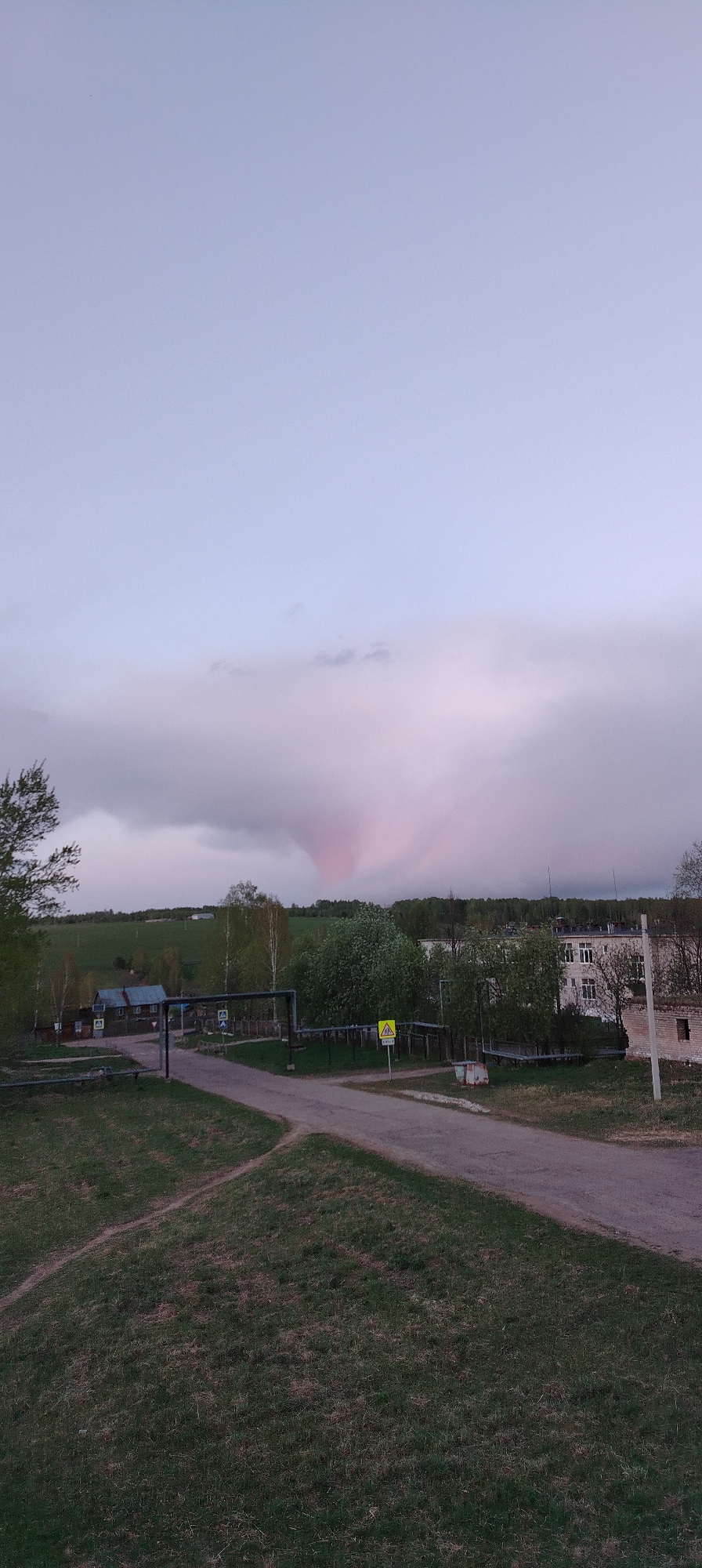 The image shows a view in Zipunovo, Russia, with a predominantly gray sky. In the sky, there is a distinct pink-tinted cloud formation that resembles a funnel shape. The foreground features a grassy area leading to a paved road with a fork in it. A series of pipes and metal structures run along the side of the road. Vegetation, including trees and shrubs, are visible in the midground, along with houses and buildings. A yellow triangular warning sign is present near the road. The scene appears to be taken during twilight or a cloudy day.