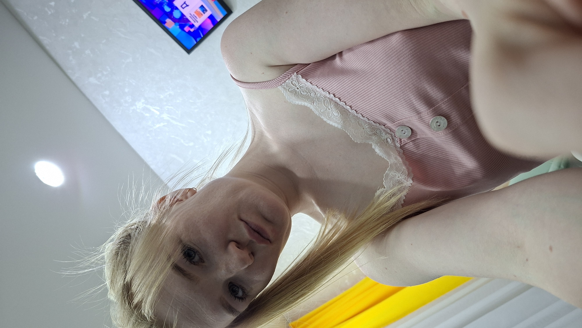 A woman with blonde hair is wearing a pink ribbed tank top with white lace trim. The scene appears to be indoors, with a television mounted on the wall in the background, displaying a colorful abstract image. A light fixture is visible on the ceiling. The perspective suggests the photo was taken from a low angle, looking upwards at the woman. There is a hint of yellow and green fabric in the lower portion of the image. The photograph was taken indoors, but there is no mention of the location in the city of Chaikovsky, Russia.