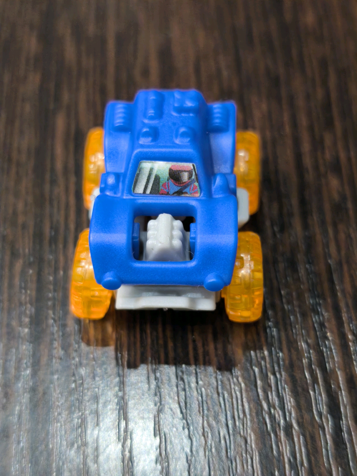 A blue toy monster truck with transparent orange wheels sits on a dark brown wooden surface. The truck has a grey engine detail visible through the front grille and a sticker on the windshield depicting a driver with a red helmet. The image is a close-up, high-angle shot, with the truck positioned in the center. The environment is likely indoors, given the lighting and the wooden table. There is no information to suggest the image is located in Karachayevsk, Russia.