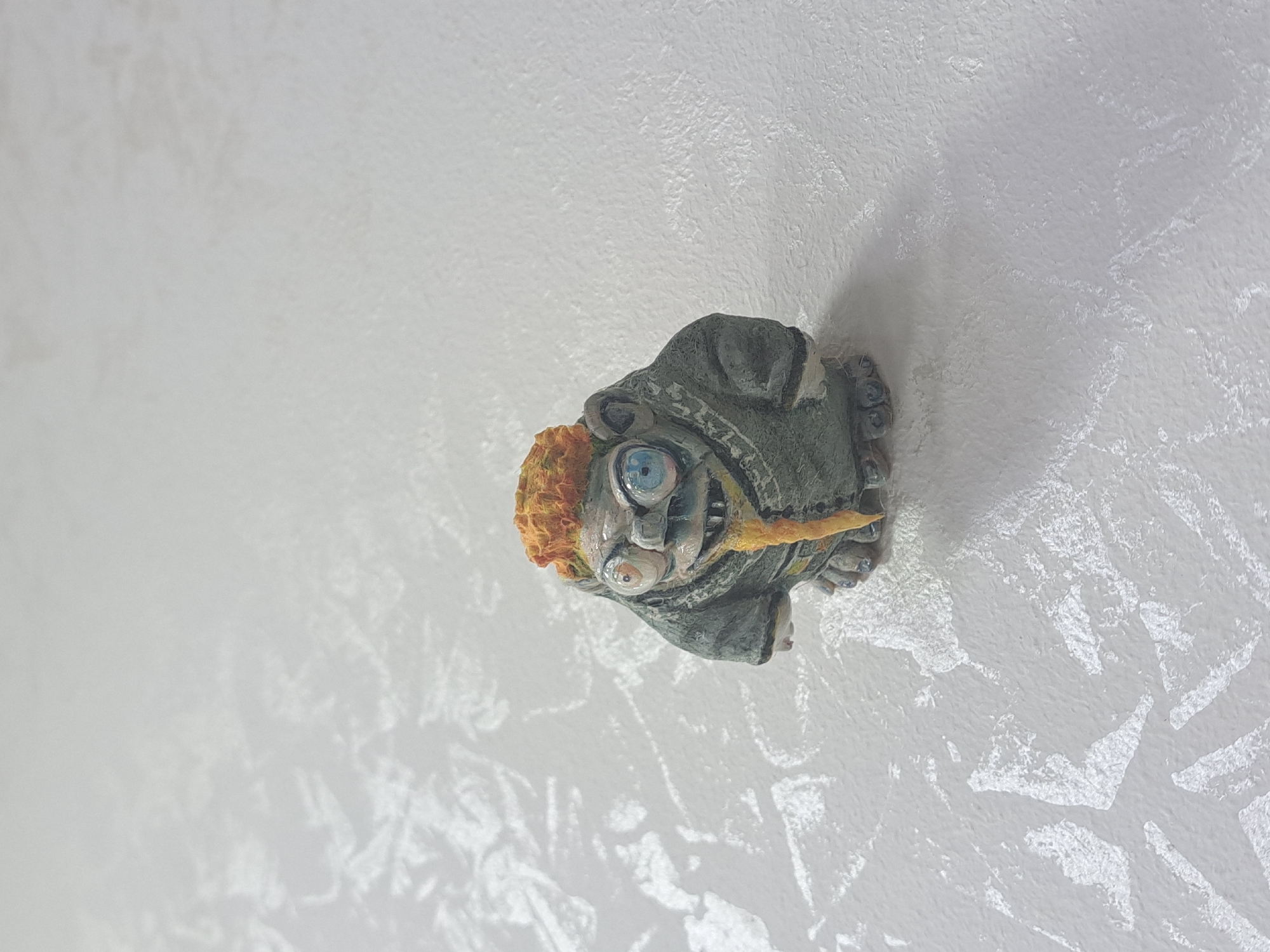 The image shows a handmade figurine lying on a white surface. The figurine depicts a whimsical character with orange hair, one large blue eye and another smaller eye, wearing a grey or green garment. It appears to be holding a yellow stick or object in its mouth. The background is a textured white surface. The location and context within Yerevan, Armenia, are not discernible from the image.