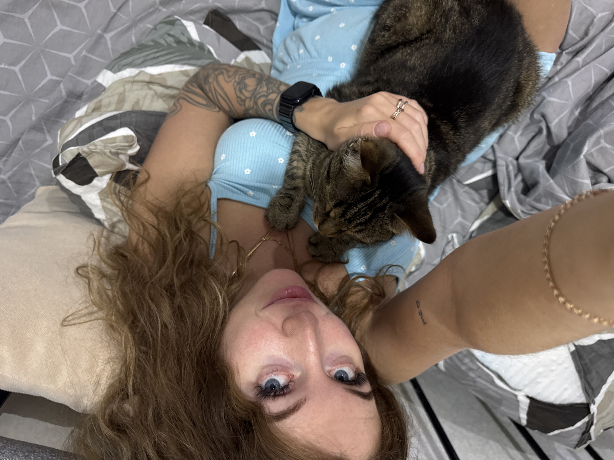 A woman is lying on a bed, possibly in a domestic setting, with a tabby cat resting on her chest. She is oriented in a head-on view, likely taking a selfie. The woman has fair skin, light brown, wavy hair, and blue eyes. She is wearing a light blue top with a pattern of small white flowers. A tattoo is visible on her right forearm, and another tattoo is on her left arm. She has a gold chain around her neck, a ring on her left hand, and a smartwatch on her left wrist. The tabby cat is resting on her chest, and her left hand rests on the cat's head. The bed has various patterns in shades of gray, white, and beige. The location is not identifiable from the image.