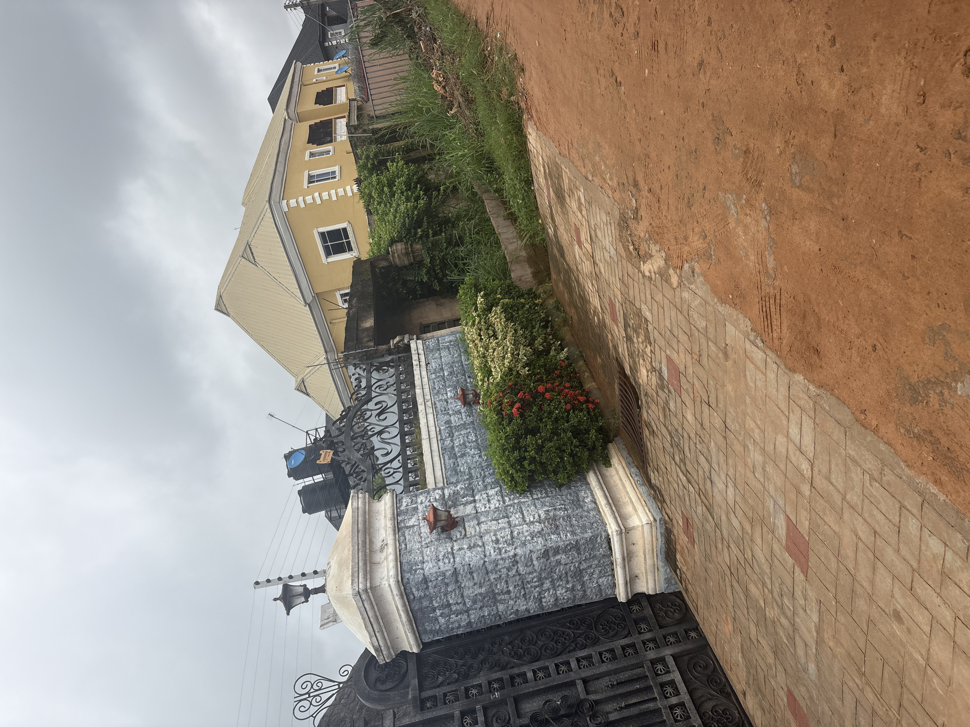 The image shows a view of a street in Obosi, Nigeria, featuring a large yellow house with a metal roof, a gray stone pillar with a lantern, a black wrought iron gate, and a paved driveway. The sky is overcast with clouds. The surrounding area includes lush green vegetation, a dirt road, and overhead electrical wires. The scene appears to be a residential area with elements of typical Nigerian architecture and landscape.