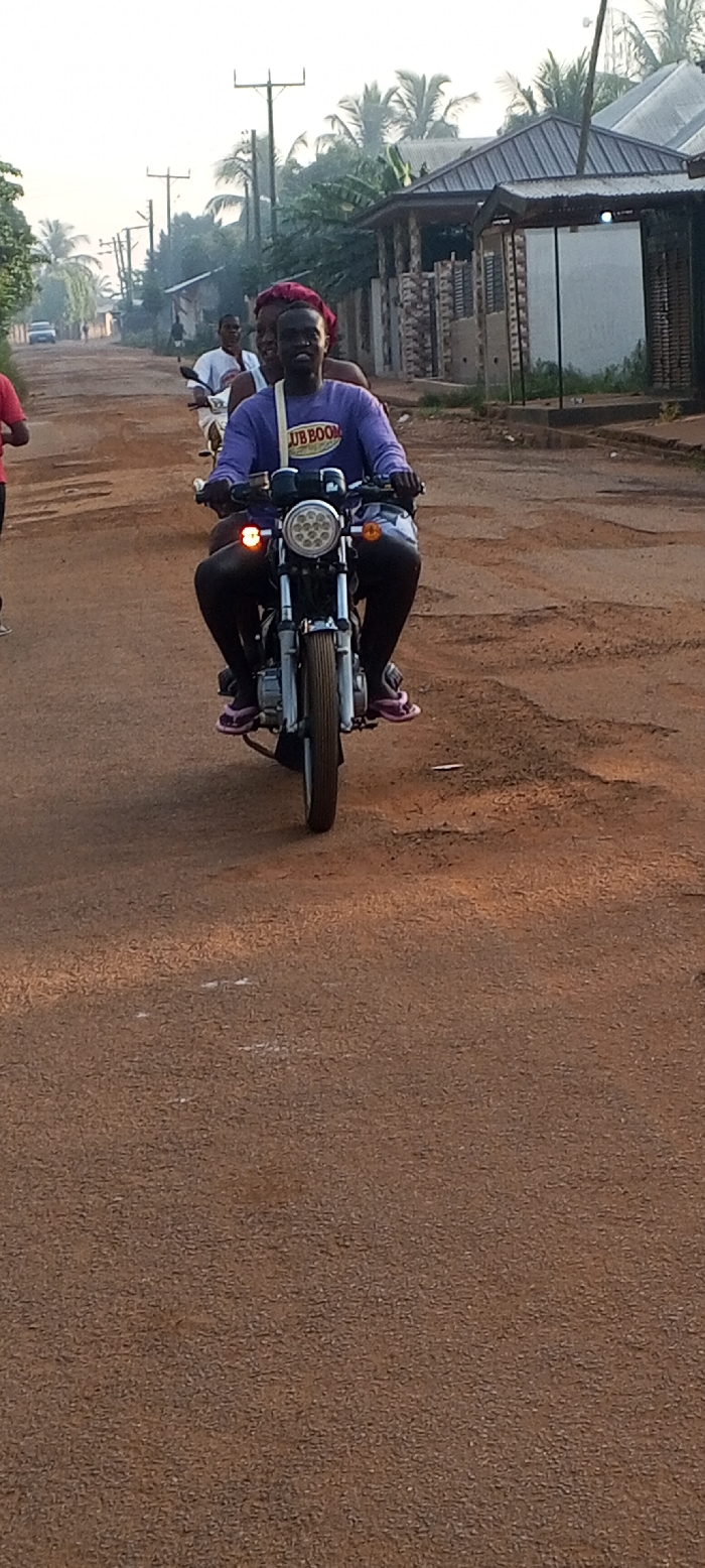 The image depicts a lively street scene on a dusty, unpaved road in Dzodze, Ghana. The soft, warm lighting suggests either early morning or late afternoon.
In the foreground, a young man is prominently featured, driving a silver and black motorcycle equipped with a circular LED headlight and amber turn signals. He wears a purple long-sleeved shirt bearing the text "JUBBOOM," dark trousers, and pink flip-flops, with a white strap or bag slung over his shoulder. He is looking towards the viewer with a broad smile. Riding behind him on the same motorcycle is another person, partially visible, wearing a red headscarf and also smiling.
The road itself is a reddish-brown dirt track, showing signs of wear with numerous potholes. Along the sides of the road, simple residential structures with corrugated metal roofs and concrete block finishes are visible. Tall wooden utility poles with overhead wires line the street, extending into the background. Lush green foliage, including several palm trees, is abundant, suggesting a tropical environment. Further down the road, other individuals are visible, including someone on another motorcycle and a person in a red shirt on the far left. The overall scene conveys a sense of daily activity and movement within a local community.