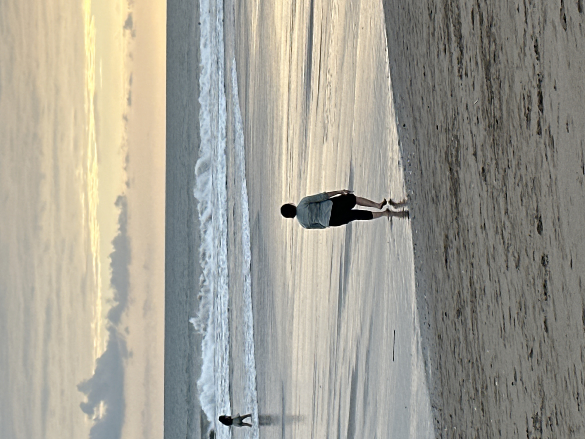 At dusk on a sandy beach in North Kuta, Indonesia, two individuals walk near the shoreline. The closer person, positioned in the foreground and facing away from the camera, is dressed in a light green top and black shorts, with their bare feet touching the shallow water. In the middle ground, a second person, also facing away, wears a dark top and light-colored shorts and is walking towards the ocean. The sea is calm with gentle waves breaking on the shore. The sky is filled with a gradient of soft yellows and grays, with scattered clouds illuminated by the setting sun. The wet sand reflects the light, creating a shimmering effect.