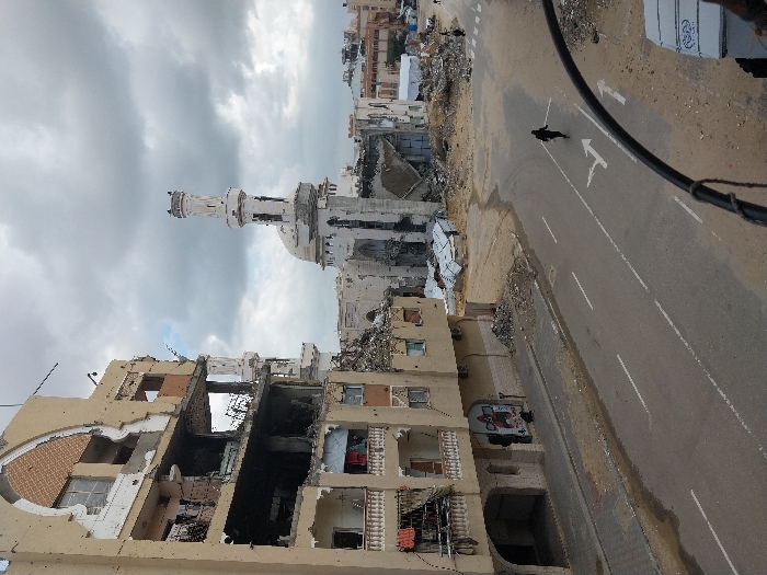 This image depicts a scene of extensive urban destruction in Khan Yunis, Palestinian Territory, under an overcast sky. In the foreground on the left, a multi-story building with a tan facade exhibits severe damage, with several floors partially collapsed, exposing interiors and rebar. Decorative arched elements on its upper section are broken, and impact marks dot its surface. Laundry is visible hanging on balconies of the lower, less damaged sections.
In the midground, a heavily damaged mosque stands, its minaret partially intact but fractured and eroded, and its main dome and surrounding structures reduced to rubble and twisted debris. Large piles of concrete and other construction materials are scattered between the damaged building and the mosque, extending into the background.
An asphalt road with white lane markings, including turn arrows, runs across the midground. One individual, clad in dark clothing, walks away from the viewer along the center of this road. Another, smaller figure is partially visible near the base of the damaged building on the left, near a wall mural. Further in the background, more destroyed buildings and mounds of debris are visible, along with a white tent structure. Black wires or cables are present in the extreme right foreground, partially obscuring the view.