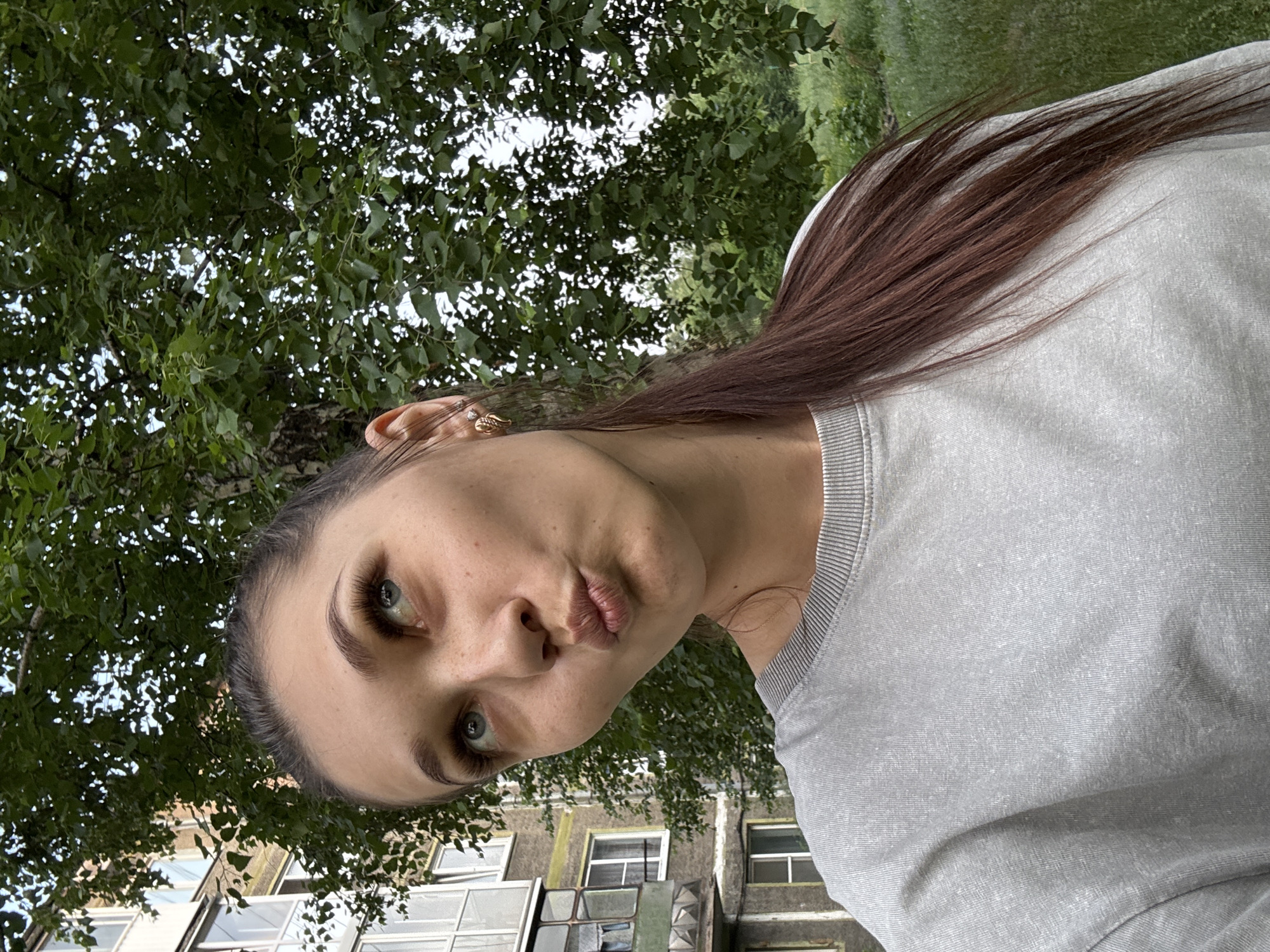A young woman with brown hair pulled back in a ponytail, wearing a grey sweatshirt, poses for a selfie outdoors. She is looking upwards with her lips pursed in a kissy face. Behind her, a multi-story apartment building with balconies is visible, along with trees and grass. The scene appears to be a typical urban residential area in Novokuznetsk, Russia.