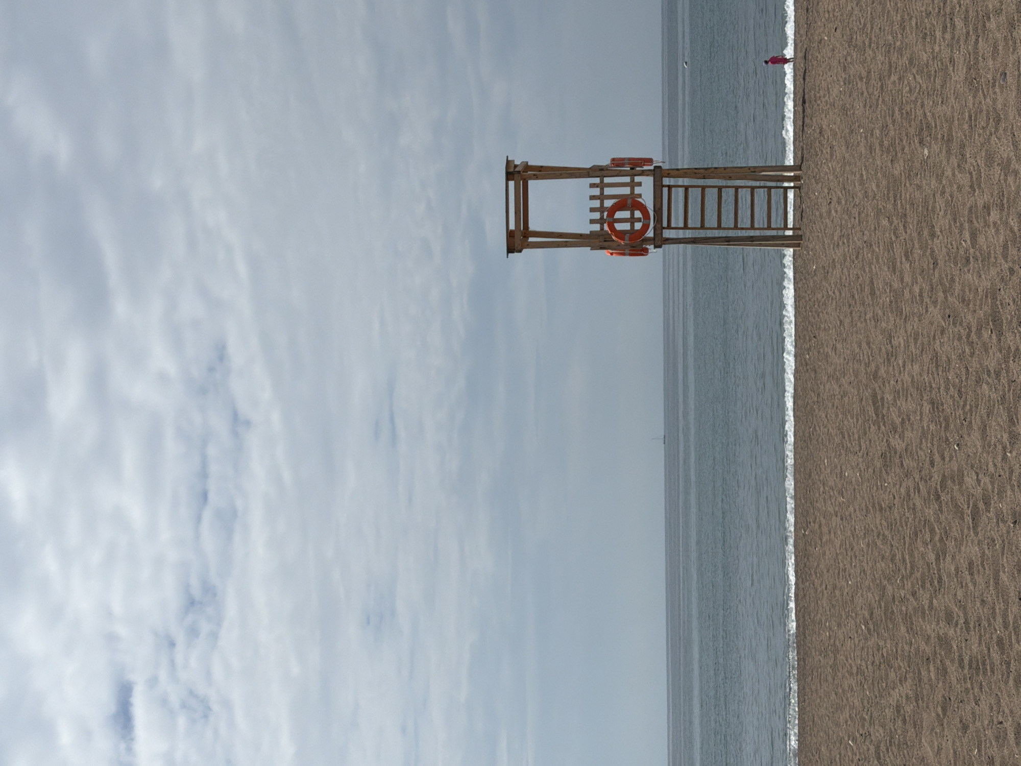 A vertical shot shows a lifeguard stand at the edge of the sea at Boreti, Montenegro. The beach is brown, and waves are lapping at the shore. A person in red stands in the distance. The sky is cloudy and gray.