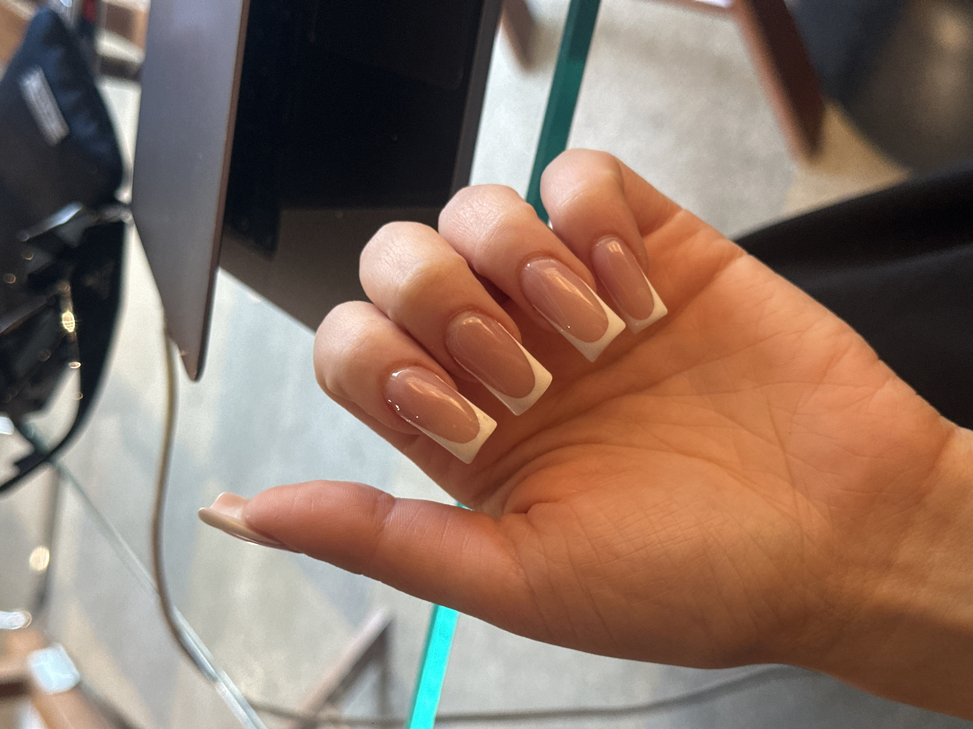 A close-up shows a woman's hand with a fresh manicure featuring long, square-shaped nails with a classic white French tip design. The hand rests on or near a clear glass surface and a turquoise object, possibly a piece of decor or part of the furniture. In the background are elements suggesting an interior setting: the corner of a laptop, what appears to be office equipment, and a blurred chair. The scene does not contain enough information to specify the location in Yerevan, Armenia.
